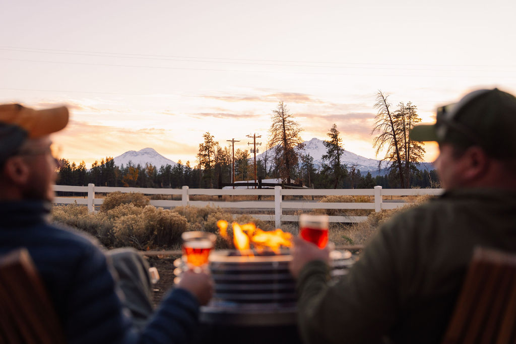 Two people sit outdoors near a fire, holding drinks, with a landscape of trees, mountains, and a sunset in the background.