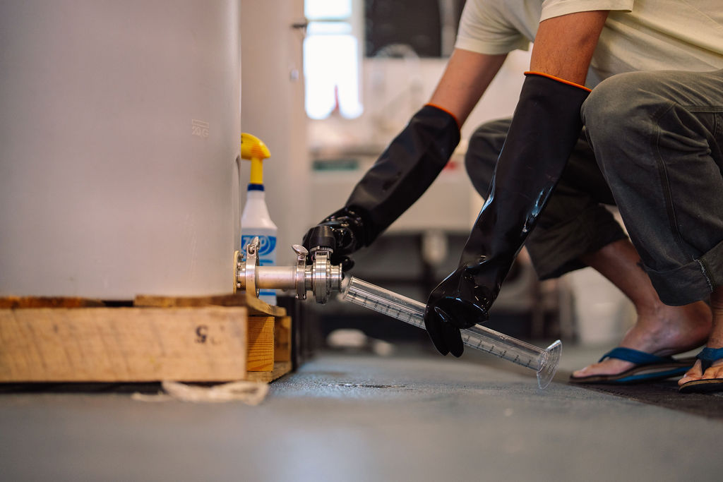 Person wearing black gloves and flip-flops, crouching near a water tap with a hose, indoors. Bright light in background.