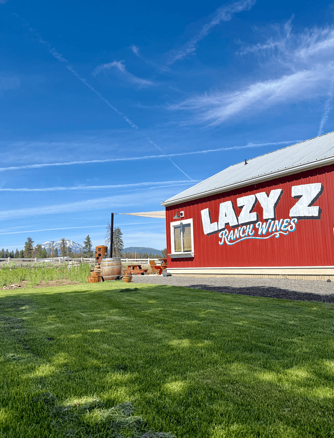 Red building with white sign reading 'Lazy Z Ranch Wines' against blue sky with clouds, green grass in foreground.