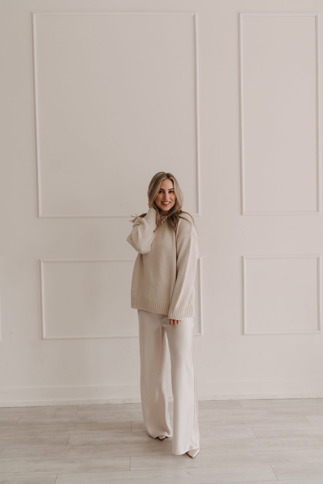 Woman with long blonde hair in beige and white outfit standing against a white paneled wall, smiling.