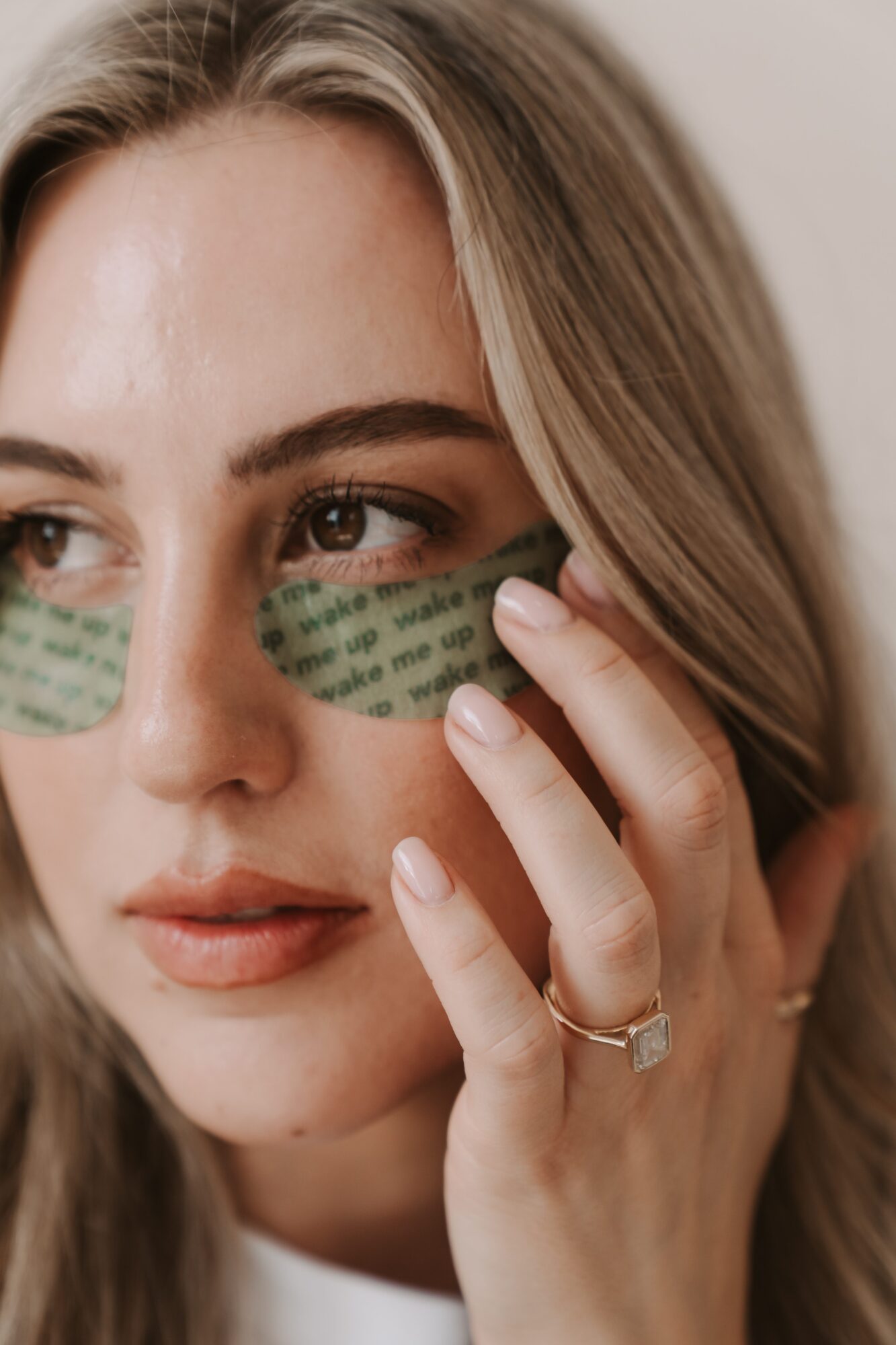 Close-up of a woman with green eye masks, touching her face, with light brown hair and a ring on her finger.