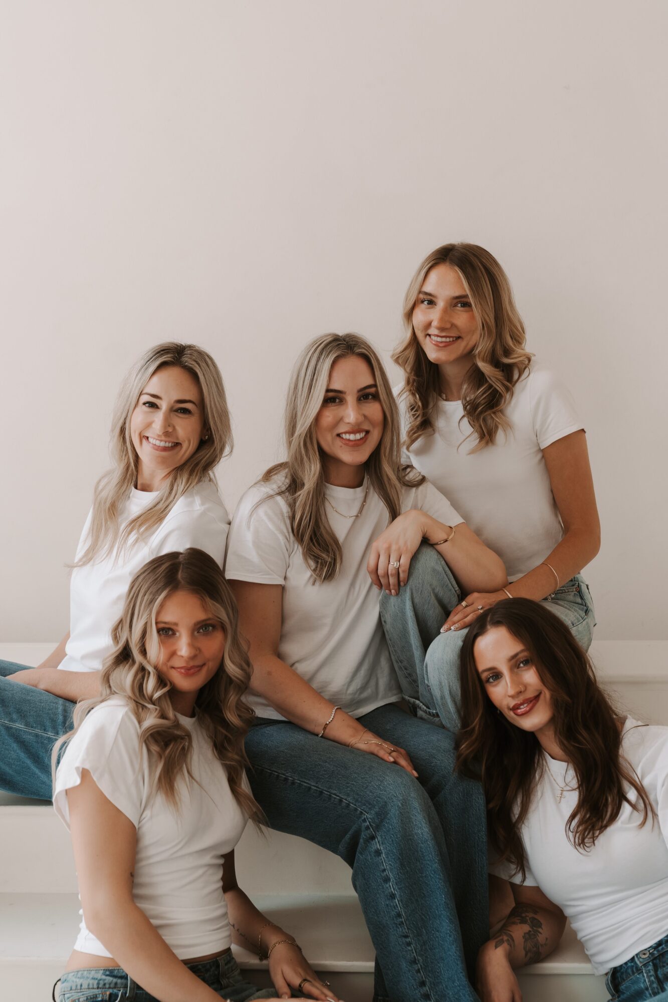 Five women with long hair wearing white shirts sitting and standing against a plain wall, smiling.