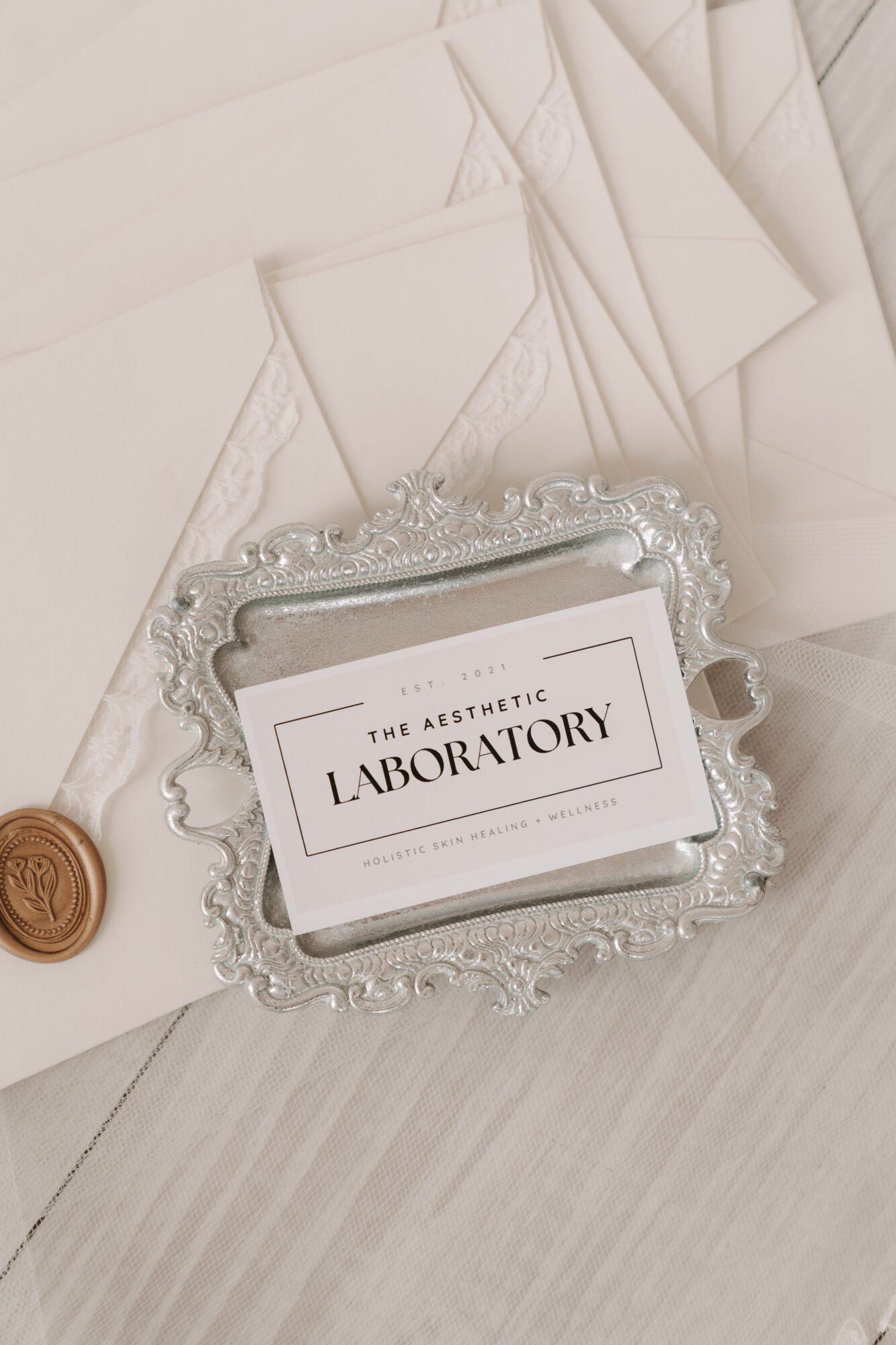 Business card with 'The Aesthetic Laboratory' on a decorative silver tray, surrounded by papers and a wax seal, on a light surface.