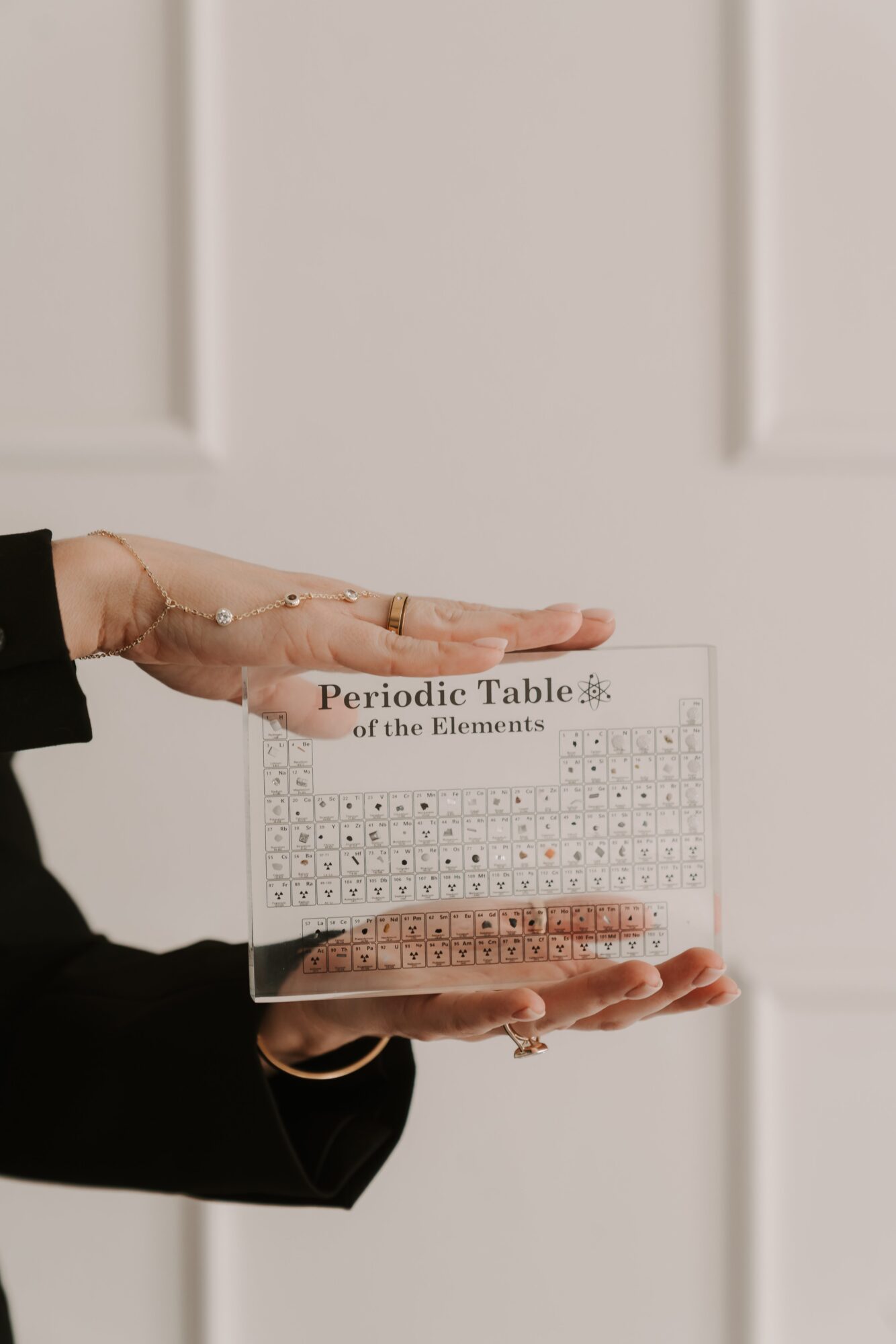 Person holding a clear rectangular object with periodic table of elements, background with white wall panels, hand with rings, black sleeve