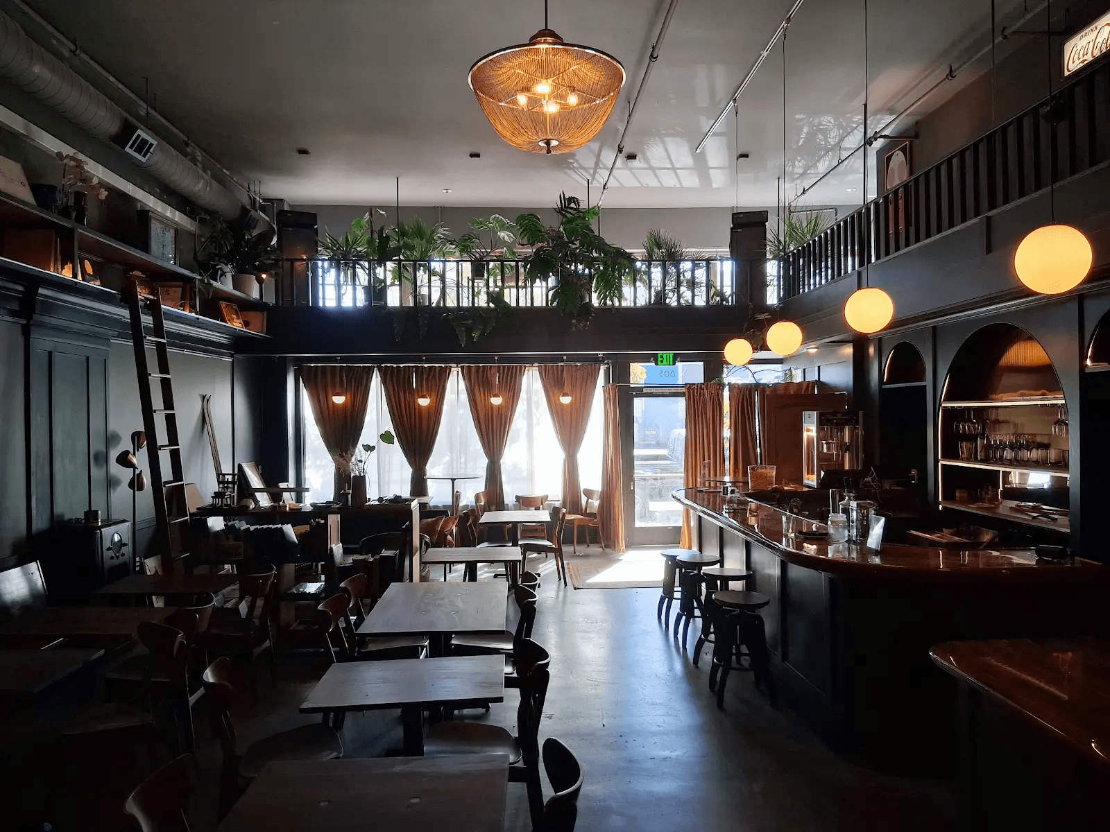 Interior of a restaurant with tables, chairs, a bar, and large windows with curtains, illuminated by hanging lights.