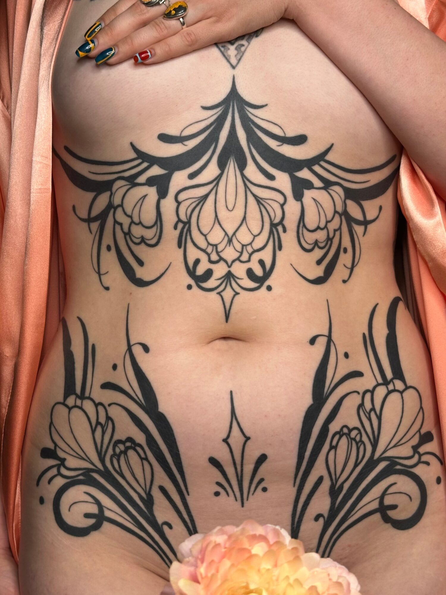 Person with large symmetrical black floral tattoos on chest and abdomen, holding their chest with hands, peach-colored fabric draped on shoulders.