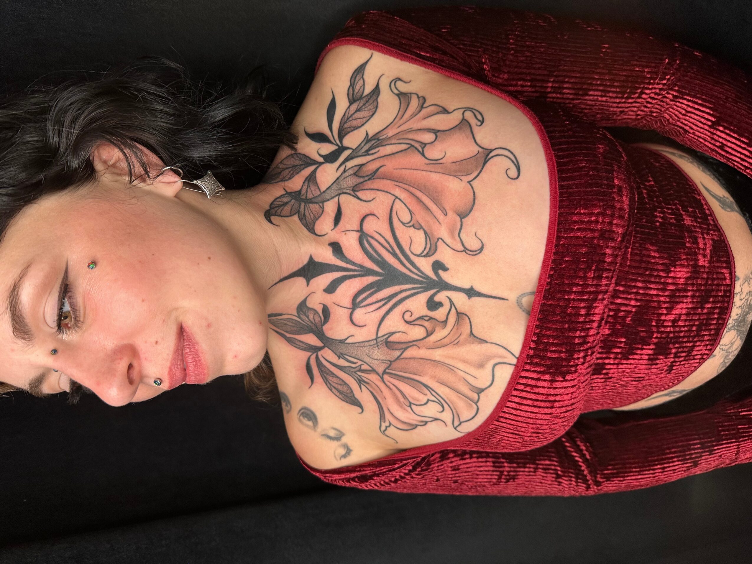 Woman with dark hair and a large floral tattoo on her chest, wearing a red velvet top, lying down.