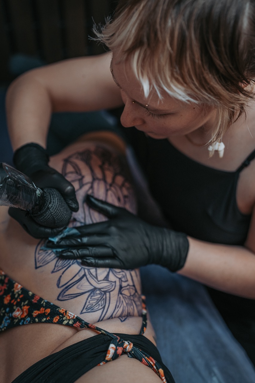 Tattoo artist in black gloves working on a detailed floral tattoo on a person's arm.