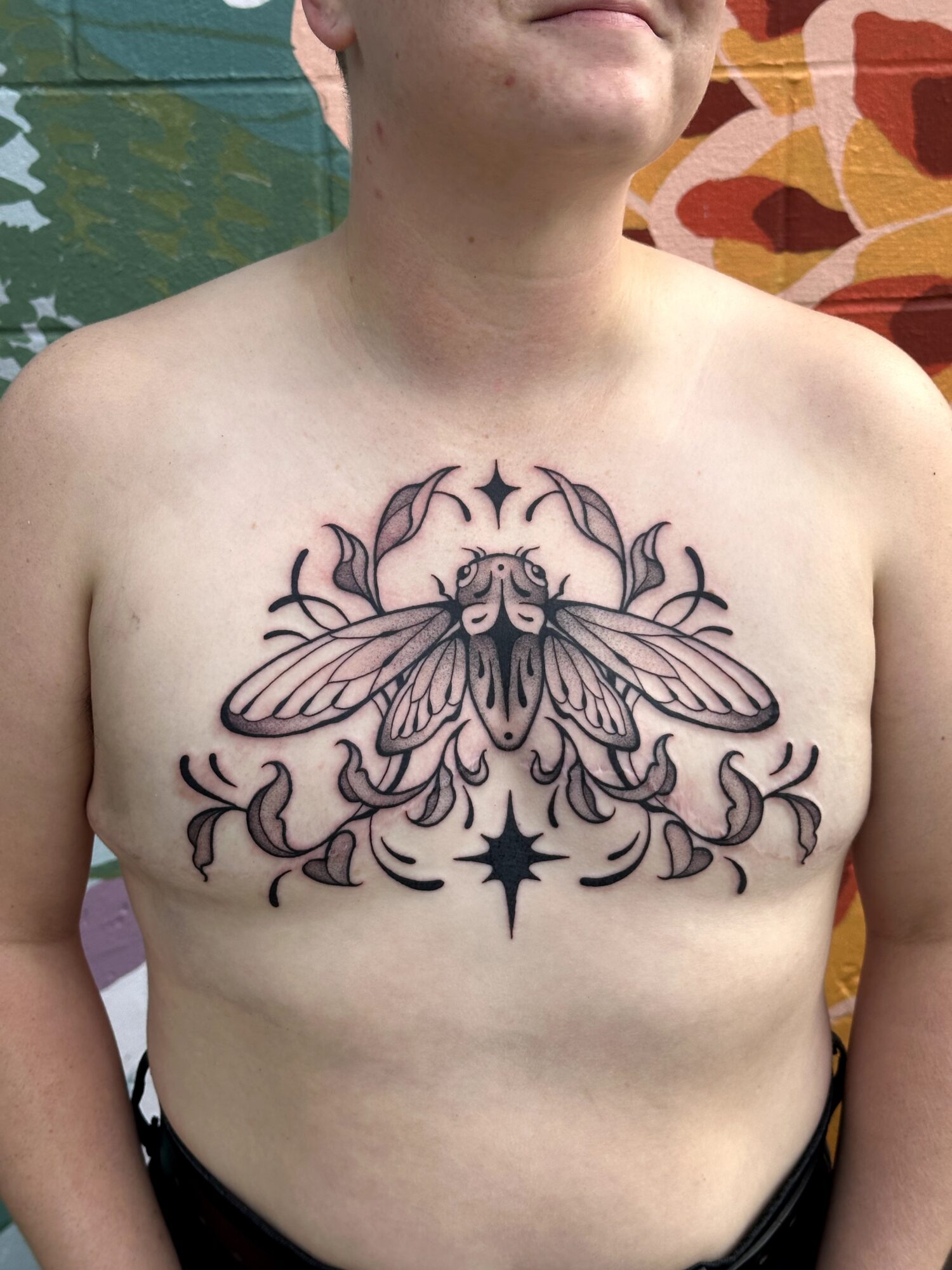 Person's chest tattoo of a large moth with symmetrical decorative elements and a star below, with colorful background.