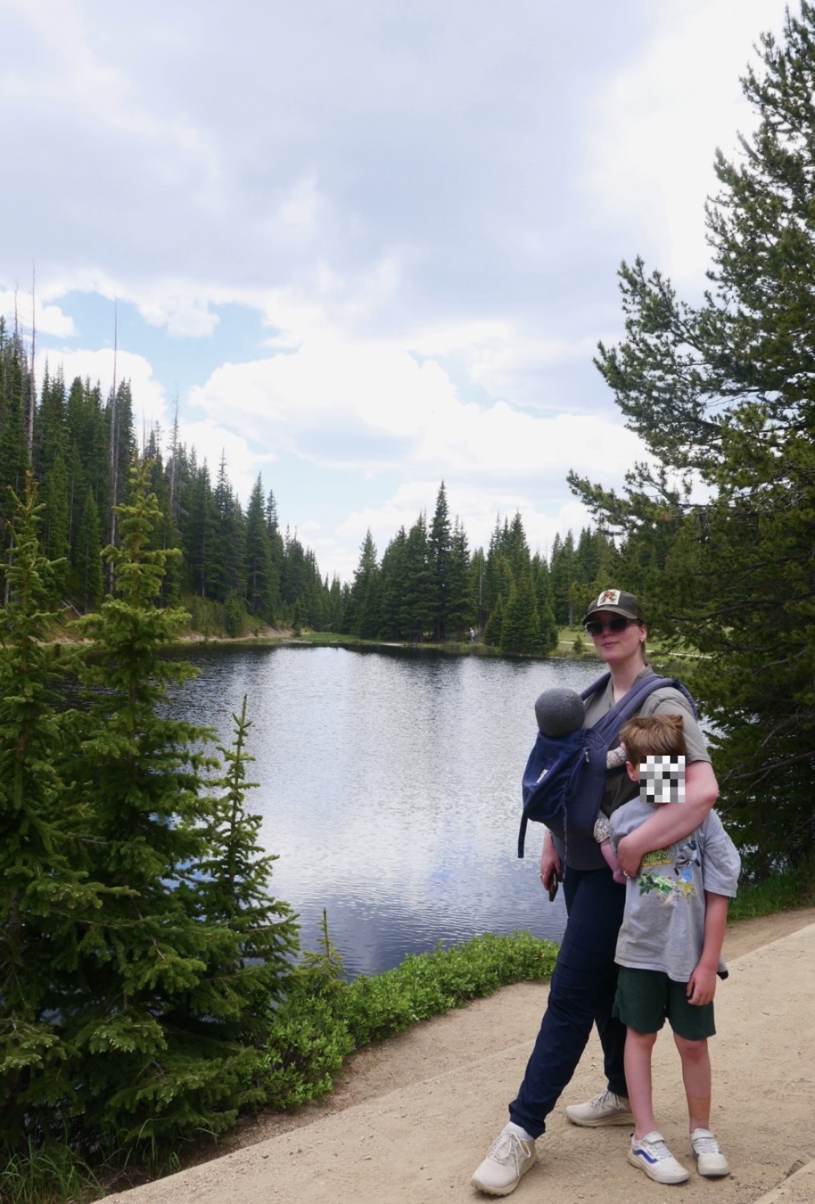 A woman and a child stand on a dirt path near a lake surrounded by trees, with a cloudy sky above.