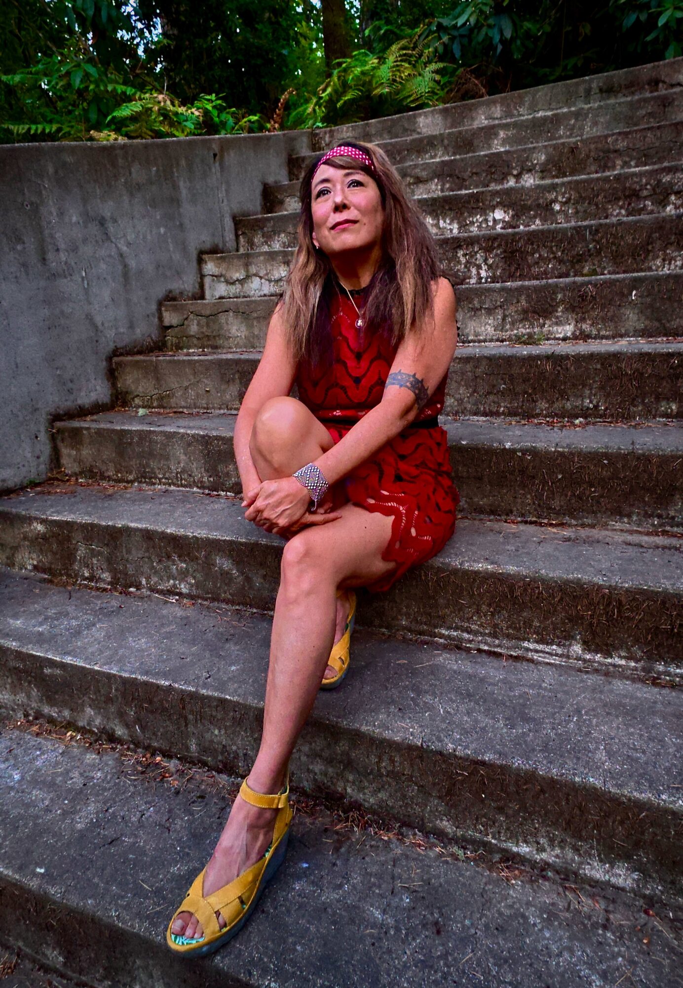 Young woman sitting on outdoor concrete stairs, wearing a red dress and yellow shoes, with greenery in background.