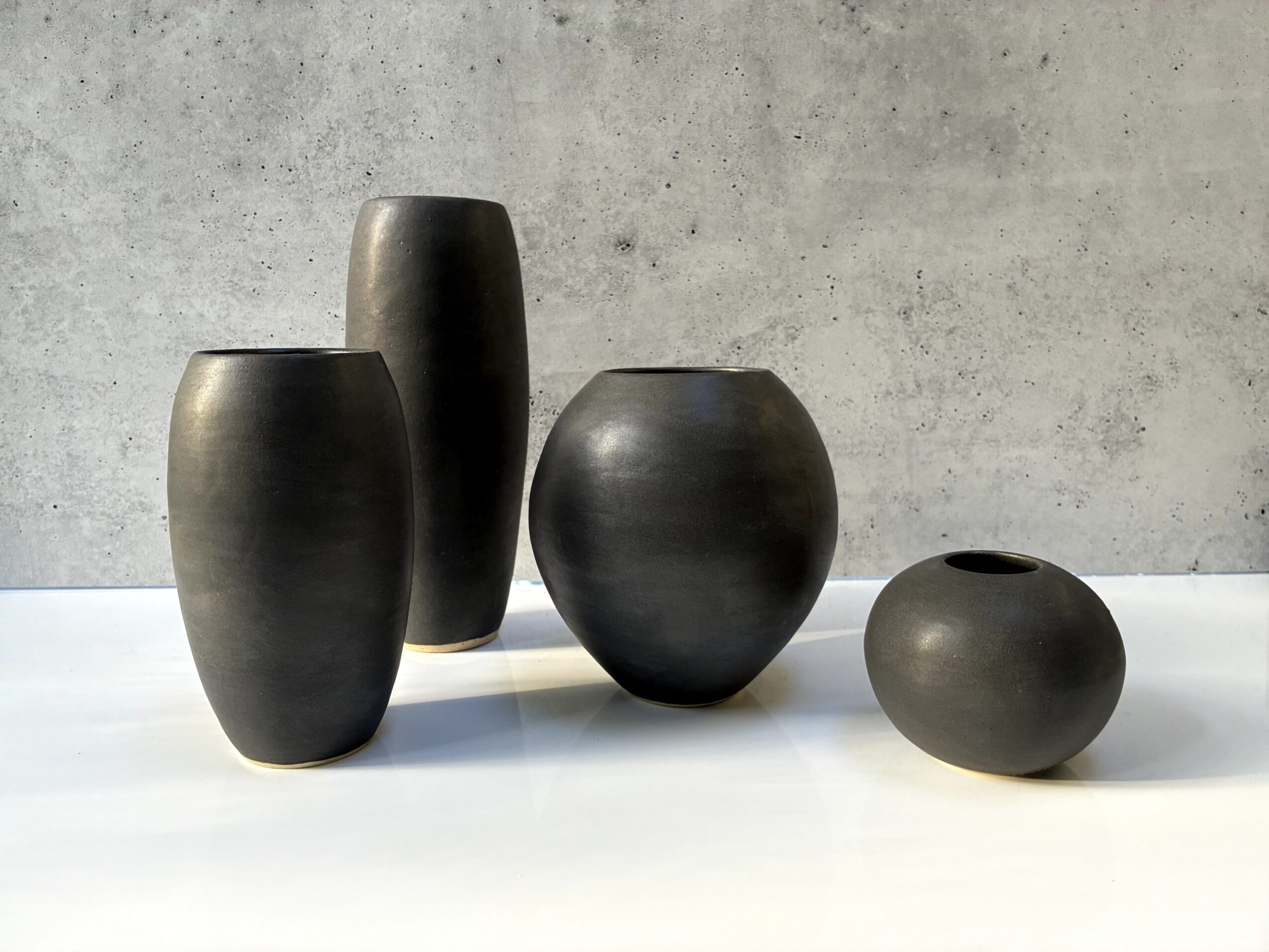 Four black vases of different shapes and sizes on a white surface against a gray wall.