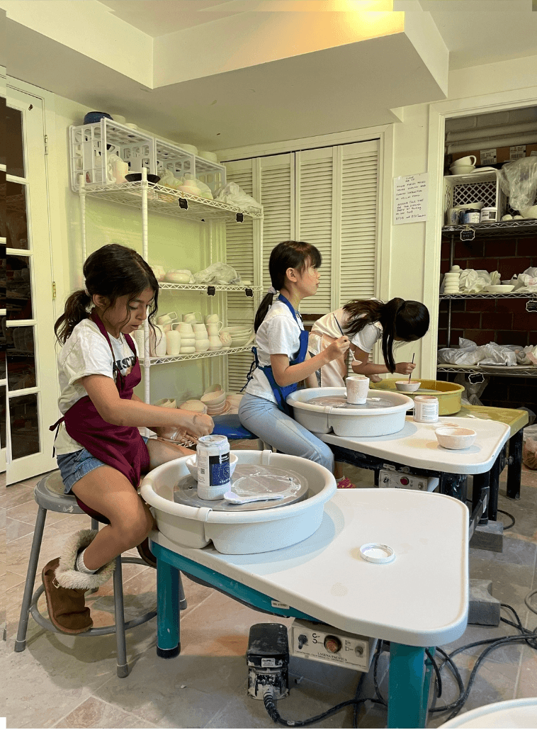Three children working on pottery projects at tables in a room with shelves and storage, focused on their clay work.