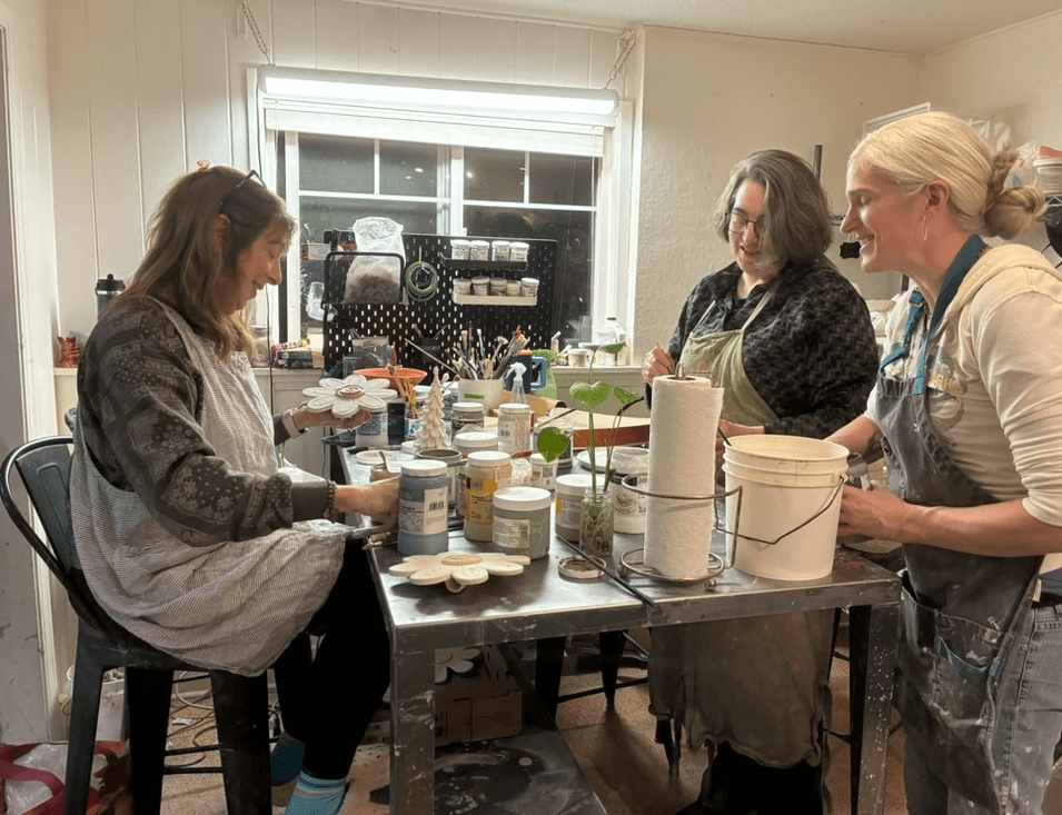 Three women working together at a table in a kitchen, surrounded by various supplies and containers.