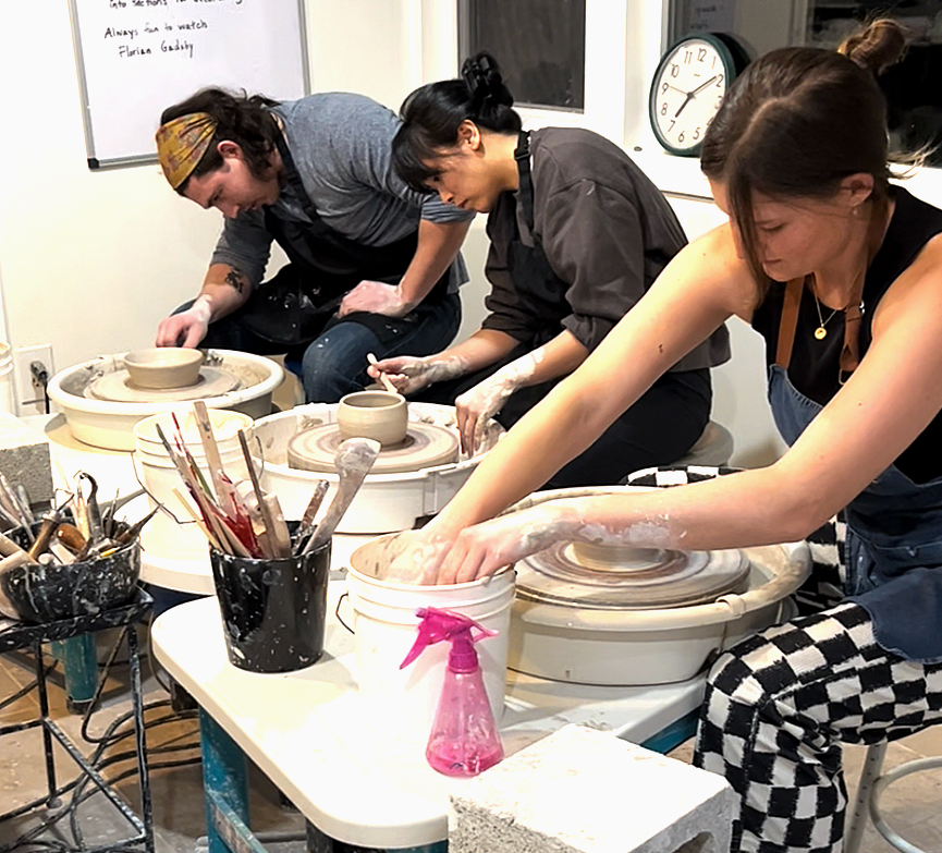 Three women are working on pottery wheels in a studio, with tools and supplies on the table nearby.