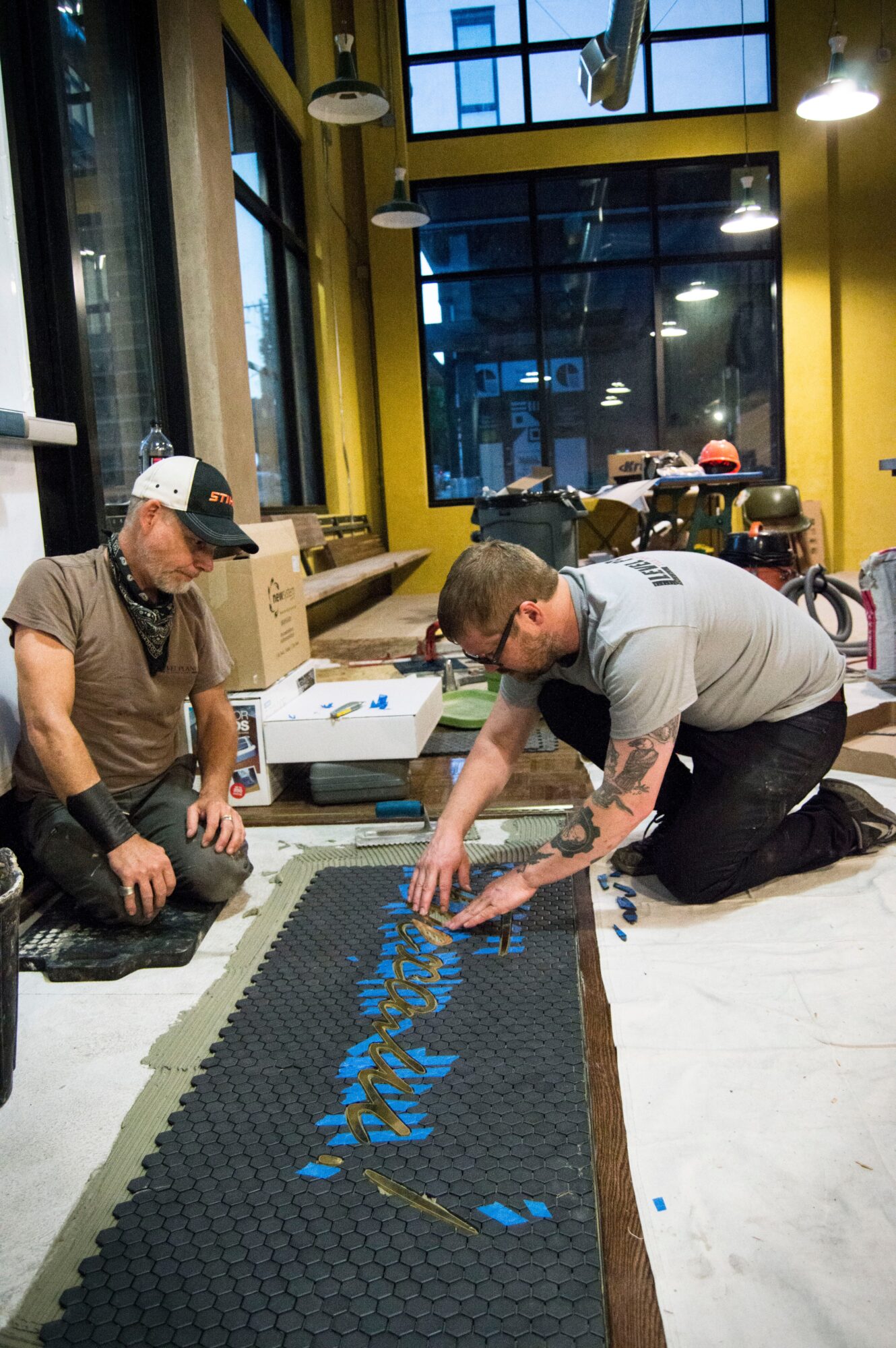 Two men working on a long black and blue textured mat inside a brightly lit room with large windows and yellow walls.