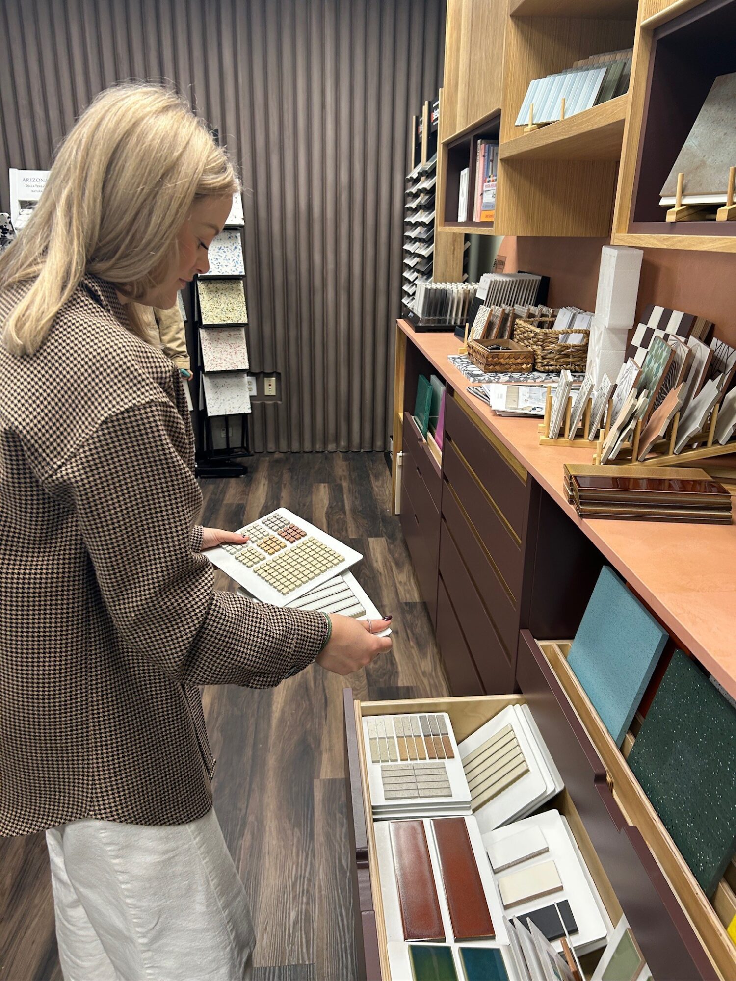 Woman browsing books in a store, shelves with books and samples behind her, wooden flooring, and a dark striped wall.