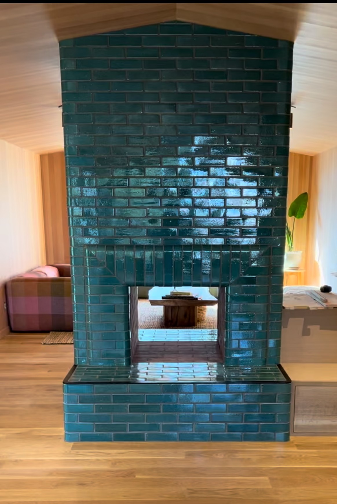 Interior view of a room with a blue tiled fireplace, wooden walls, and a wooden floor, with a sofa and window in the background.