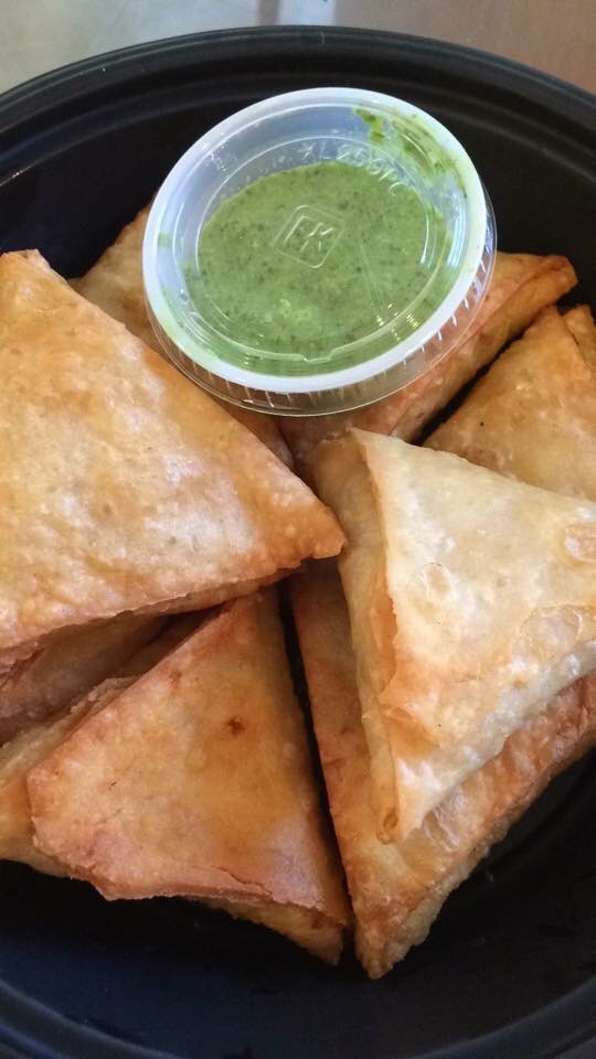Six fried triangular pastries with a small container of green dipping sauce on a black tray.