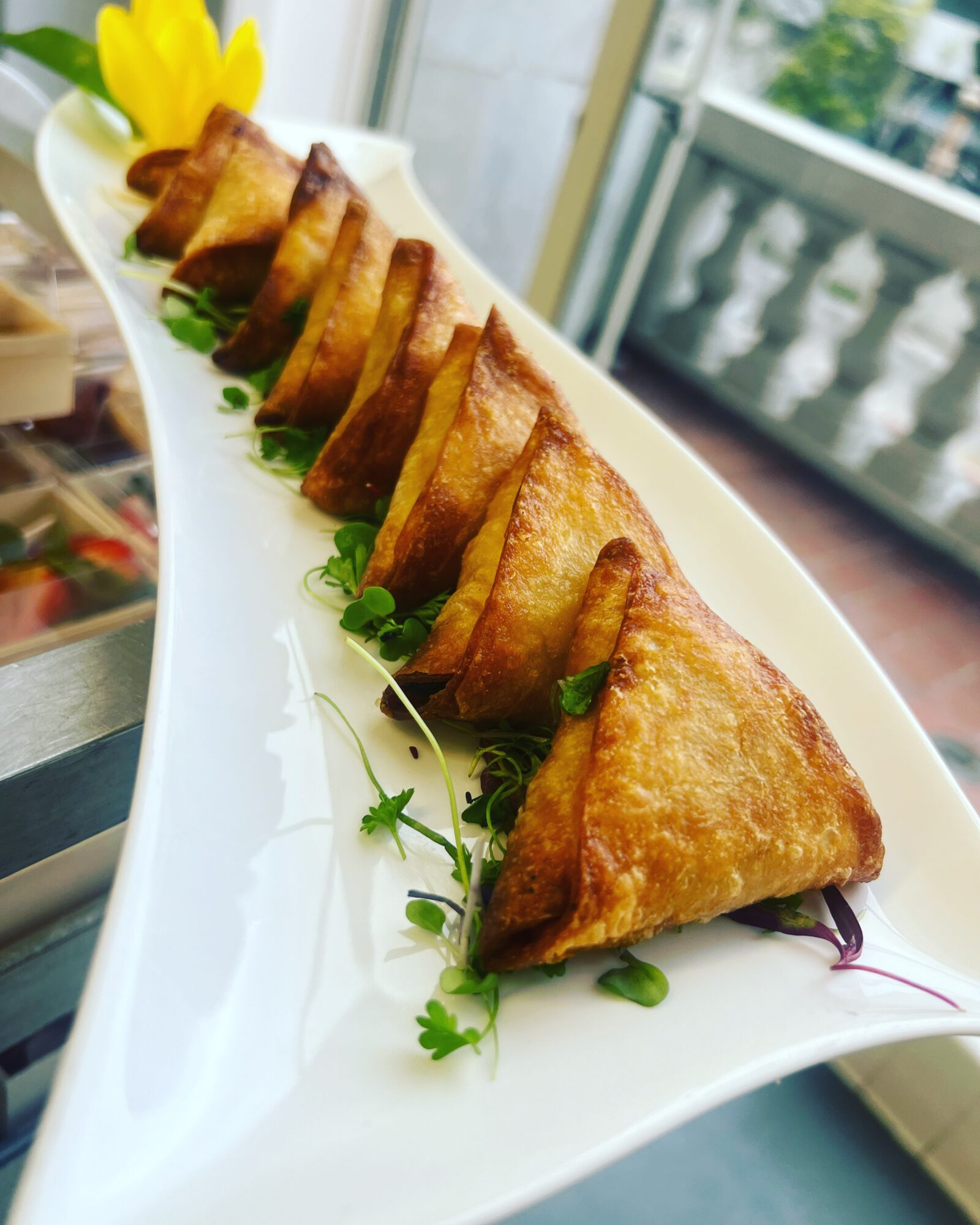 Seven fried triangular appetizers arranged on a white plate with green garnish, with a yellow flower in the background.