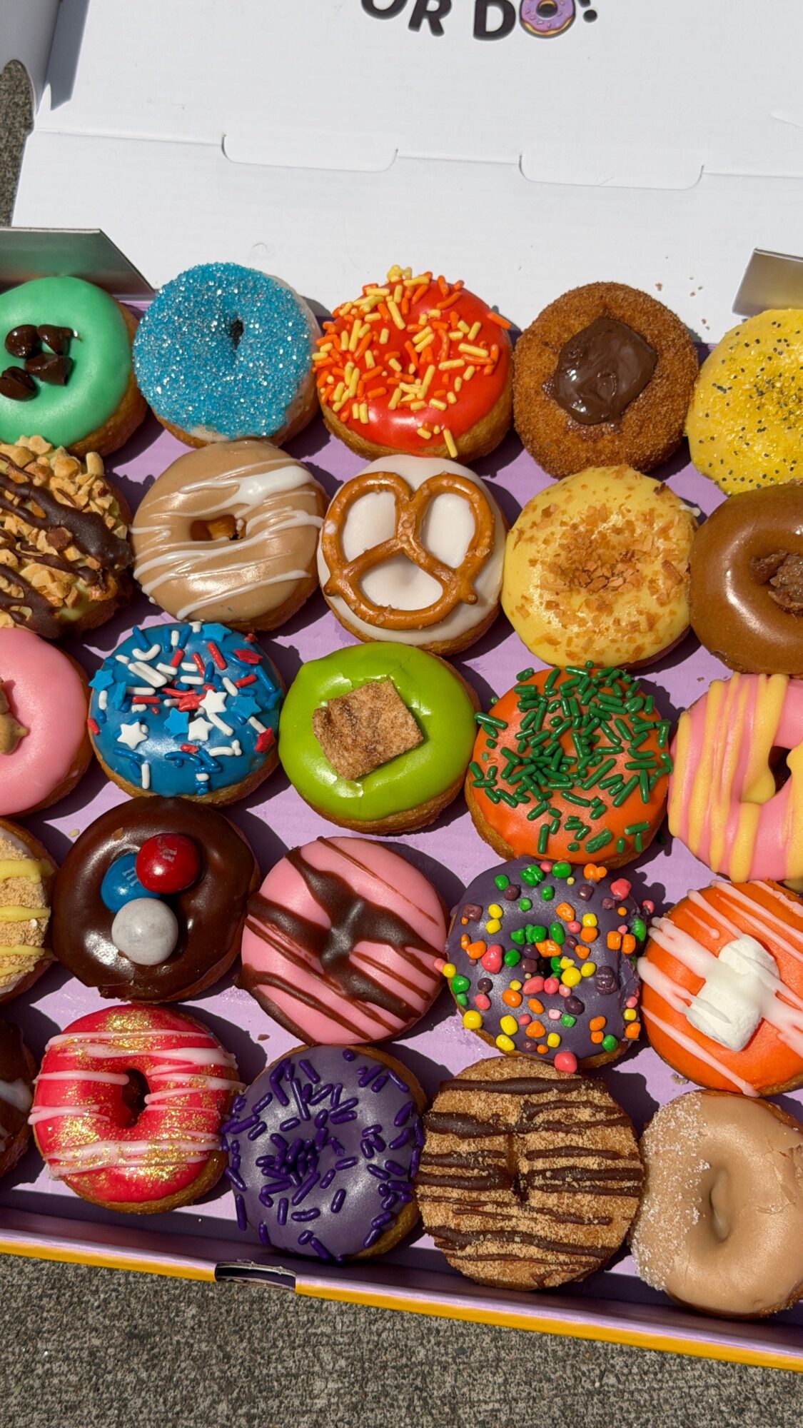 Box of assorted colorful donuts with various toppings and glazes, arranged in rows inside a box.