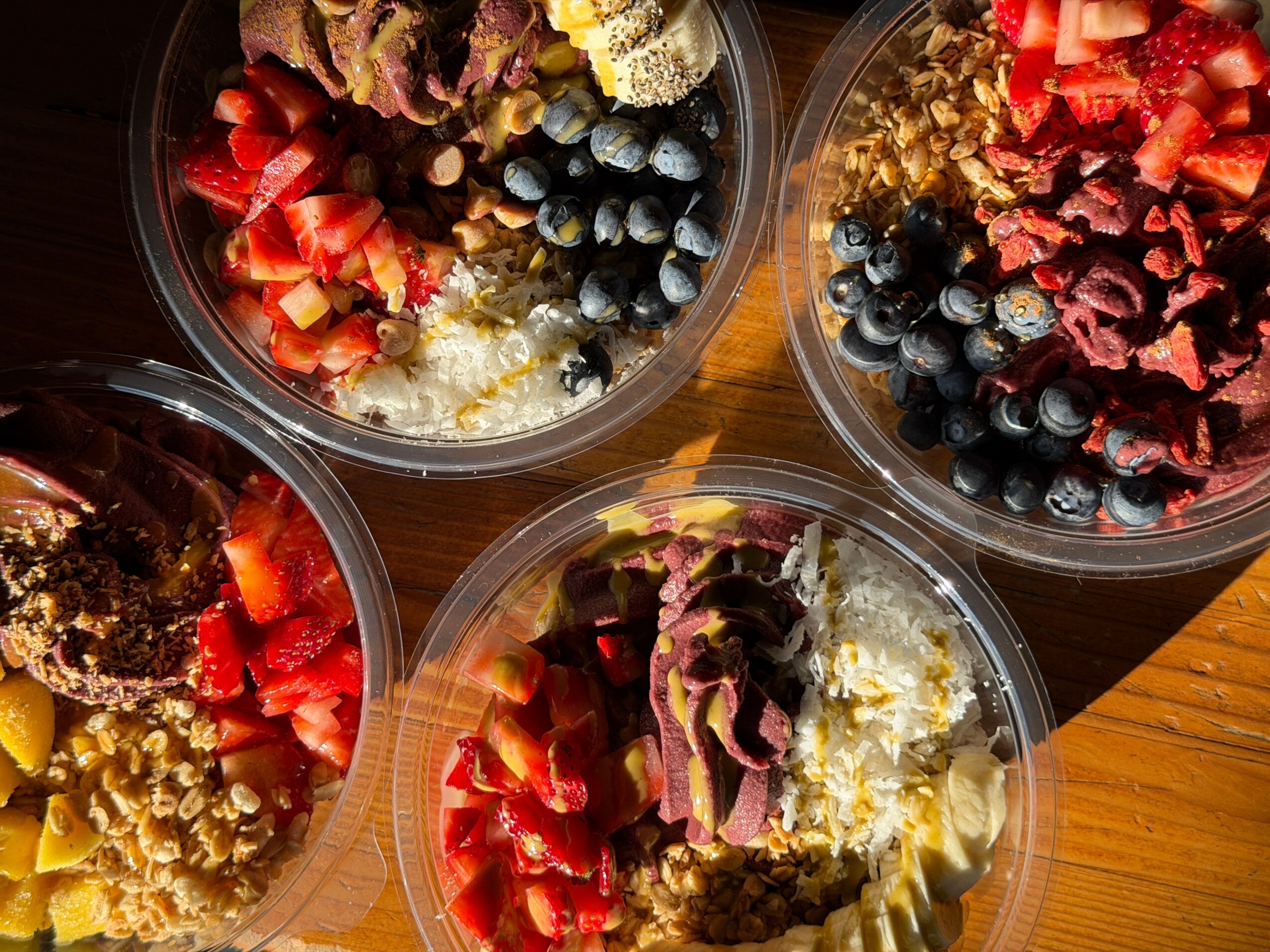 Four bowls of fruit salad with strawberries, blueberries, and other ingredients on a wooden surface.