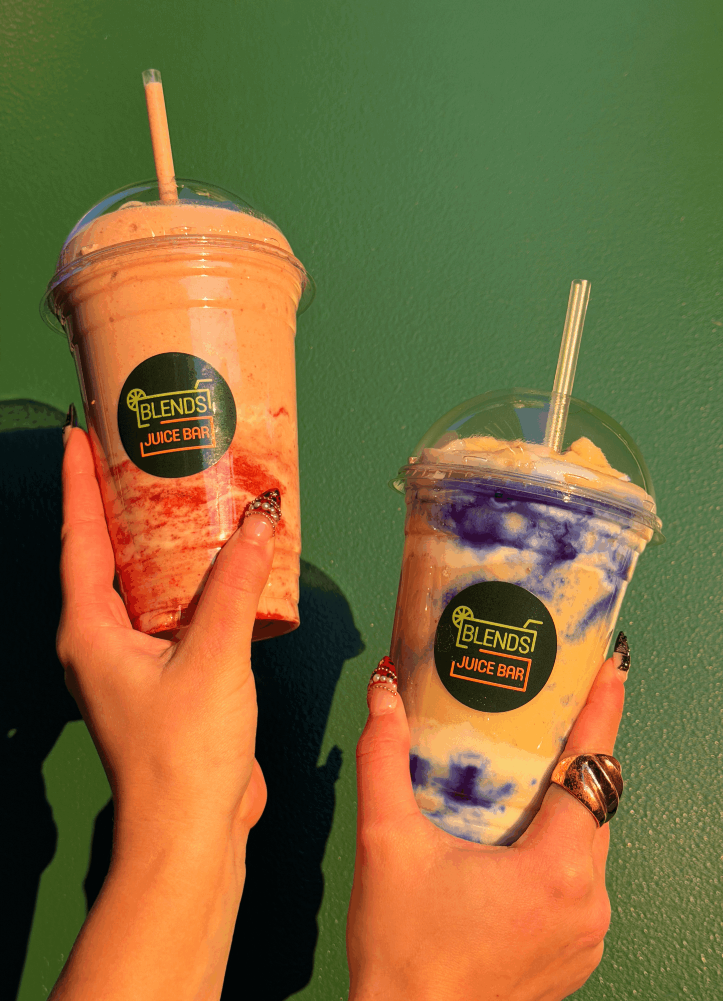 Two hands holding colorful blended drinks with straws against a green background.