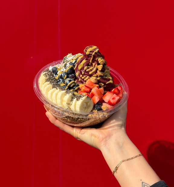 Hand holding a bowl of frozen yogurt topped with fruit, chocolate, and berries against a red background.
