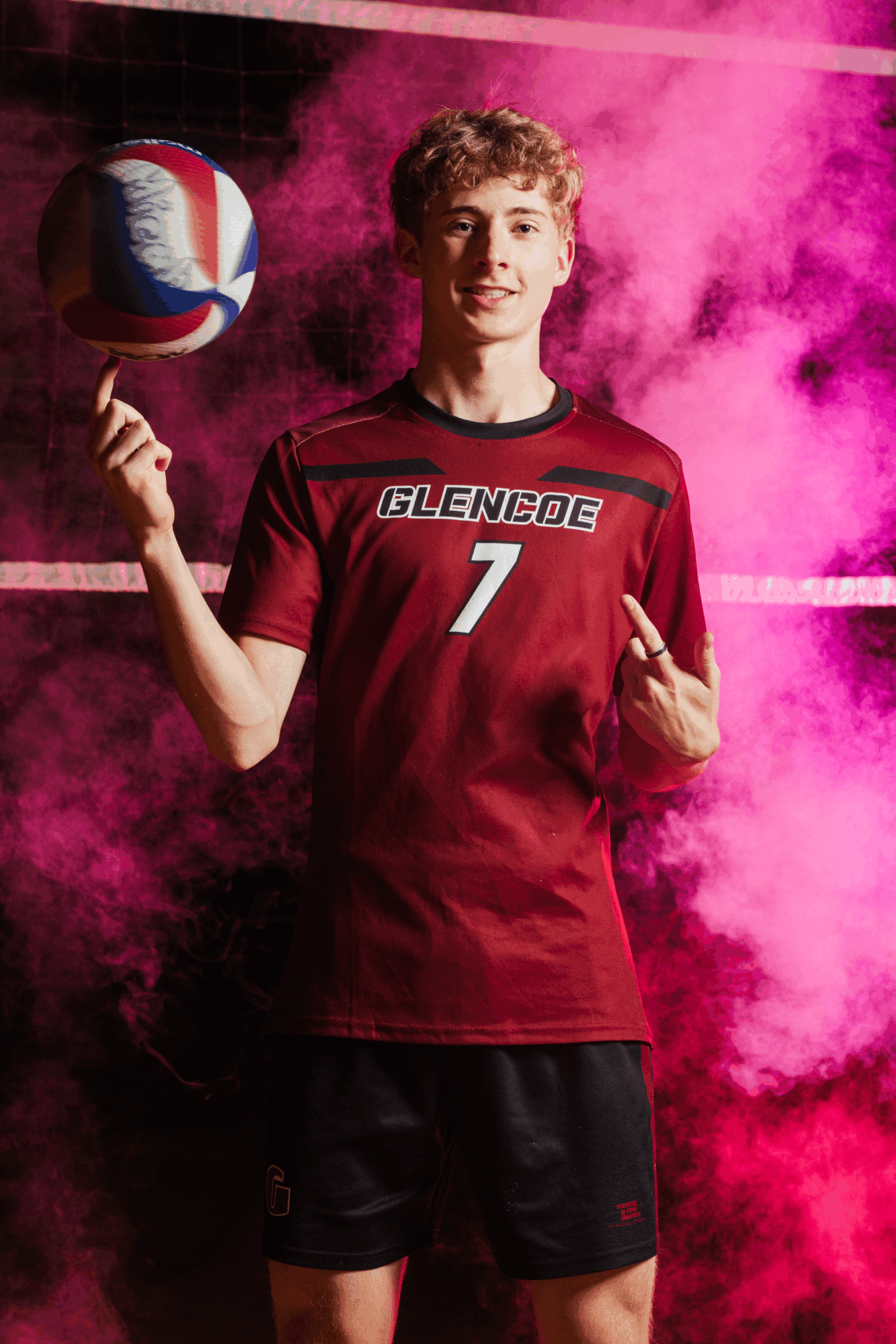 Young male volleyball player in red jersey holding a volleyball, smiling, with pink smoke background.