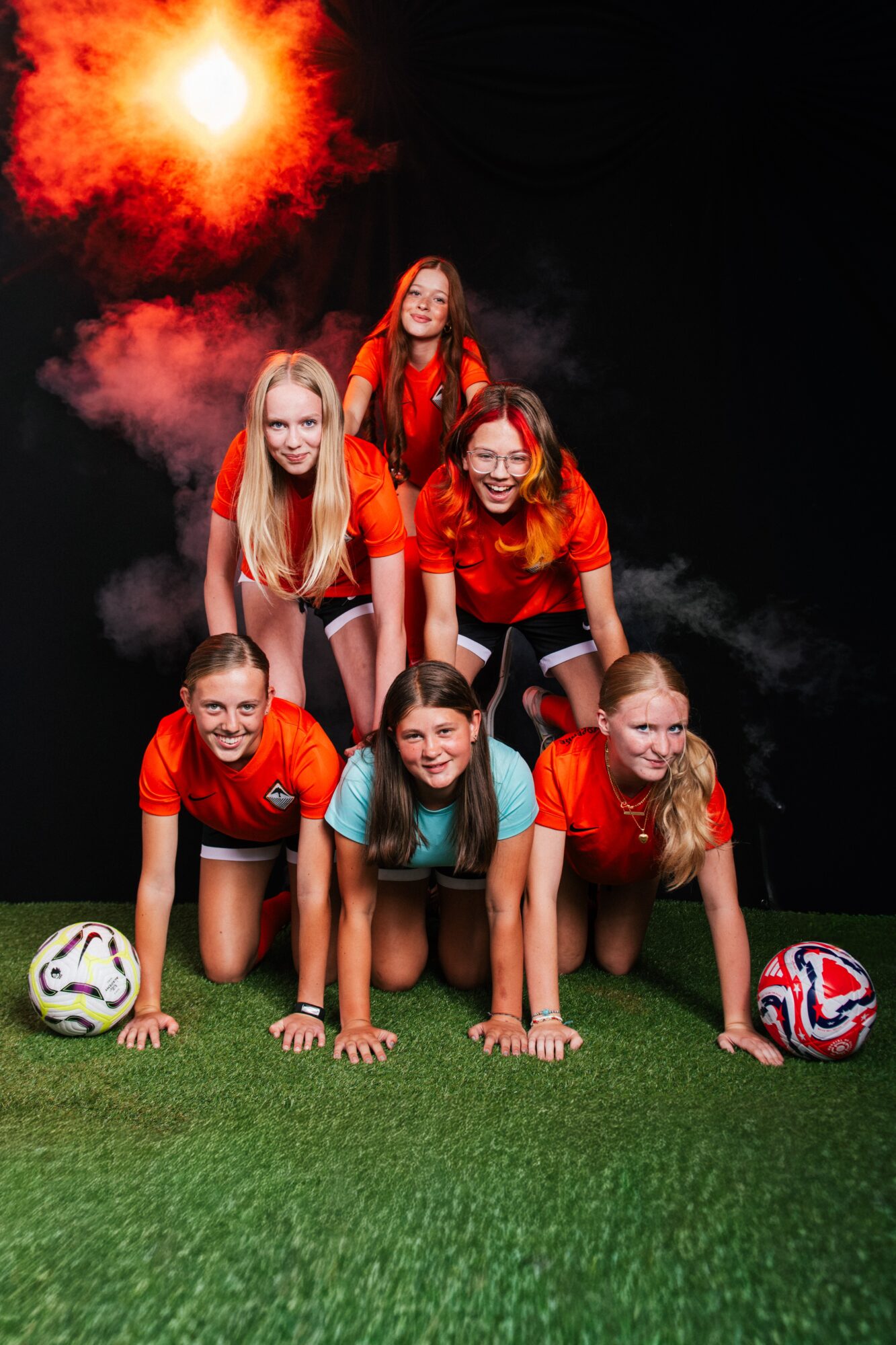Six young women in red and teal sportswear form a pyramid on a green field, with soccer balls nearby and a fiery explosion in the background.