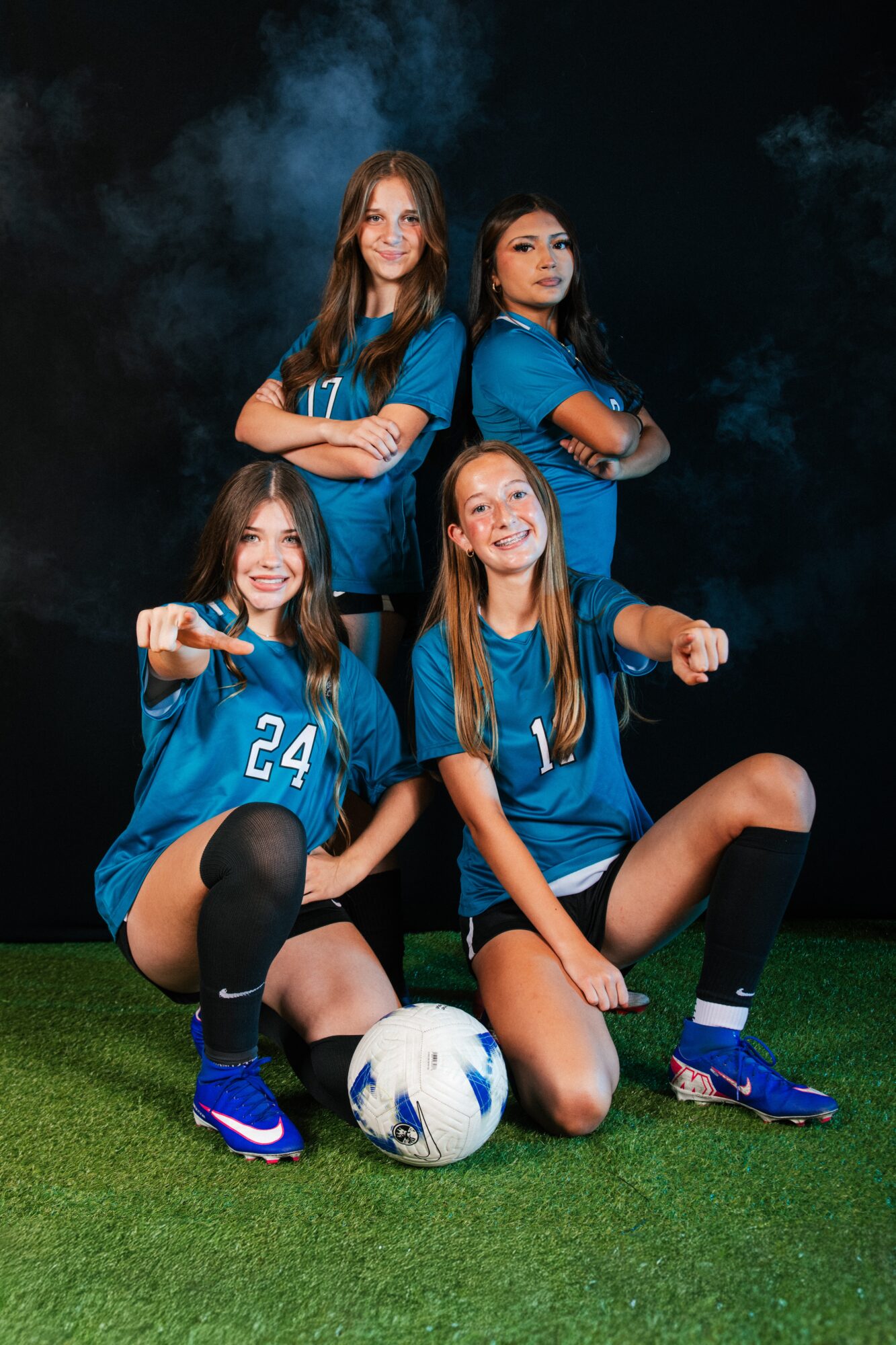 Four female soccer players in blue jerseys pose on a field with a soccer ball, two standing and two sitting, smiling.