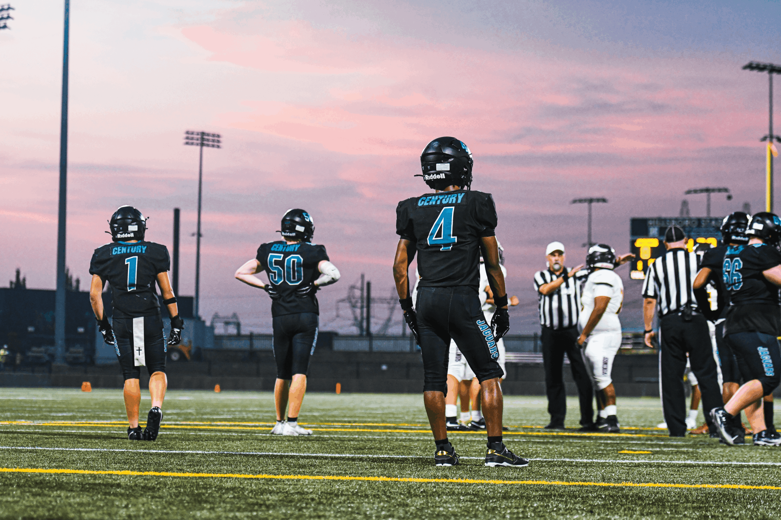 Football players on field during sunset, with referees and spectators in background, sky pink and purple, stadium lights on.