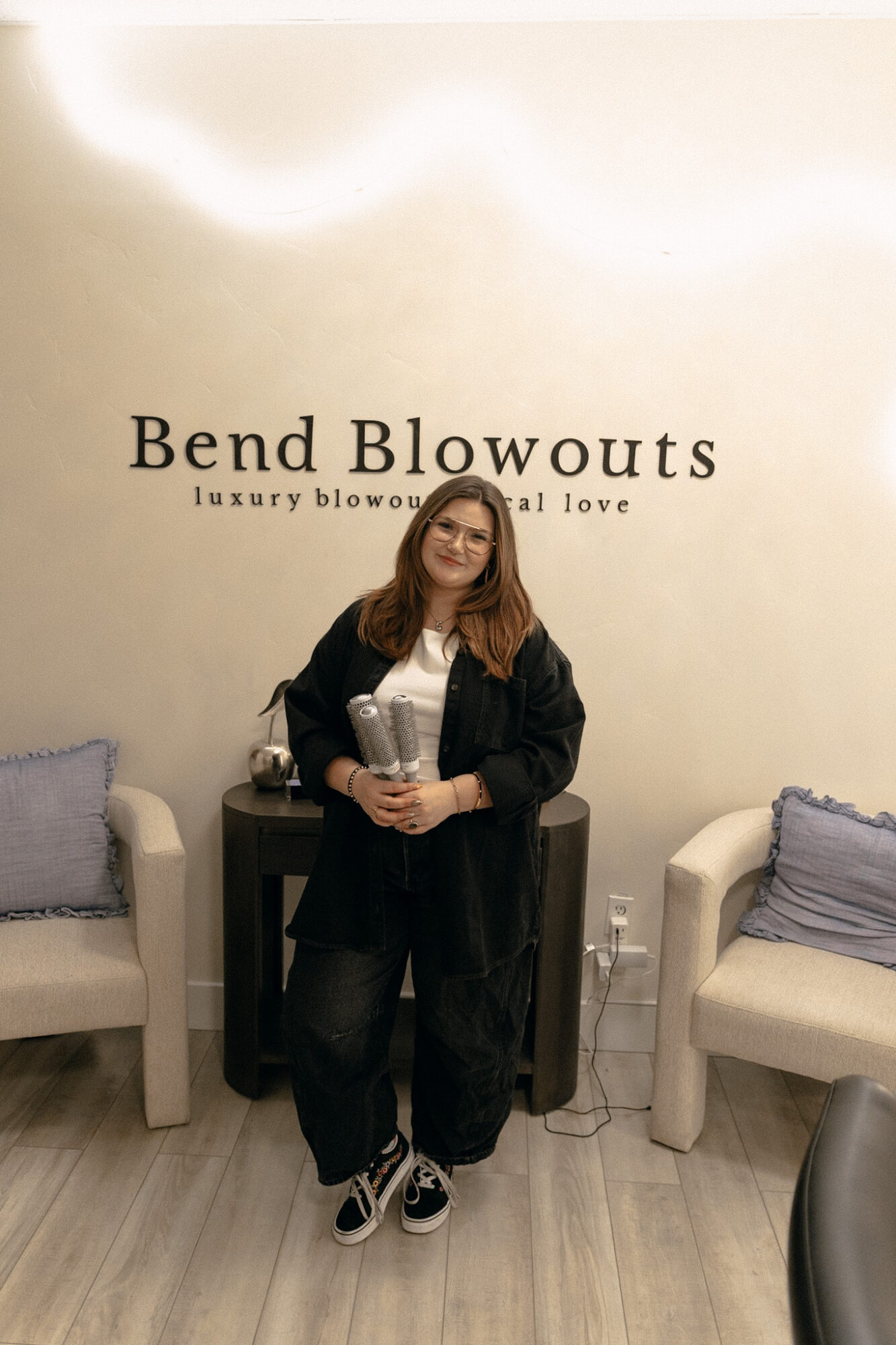 Woman standing in front of a wall with the text 'Bend Blowouts' and holding a bouquet, flanked by two chairs.