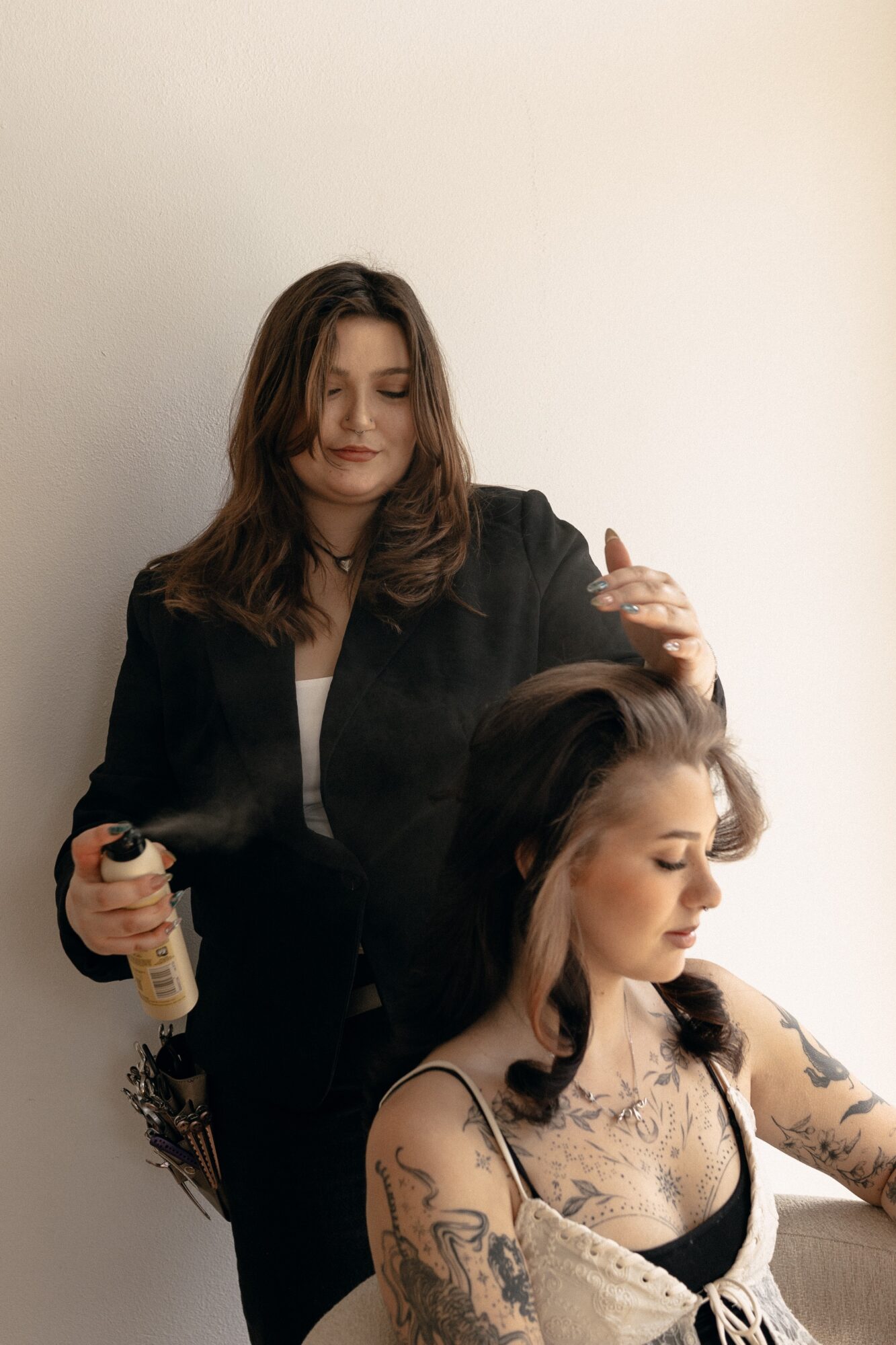 Two women, one standing and one sitting, with the standing woman styling the other's hair, against a plain wall.