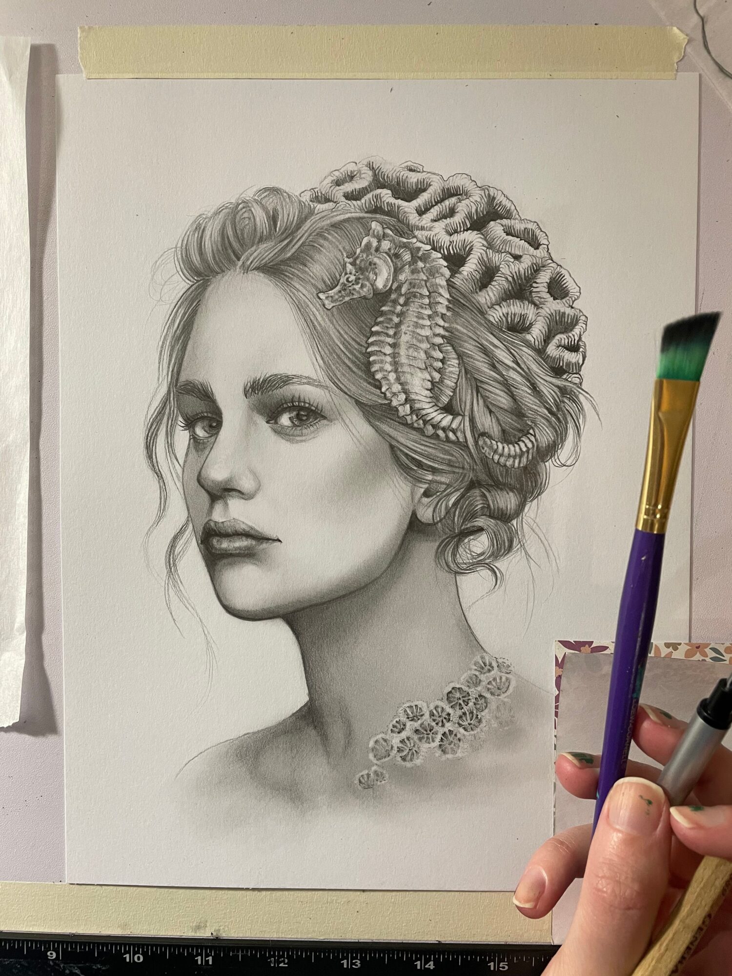 Detailed pencil drawing of a woman with intricate braided hair and jewelry, viewed from the side, with a hand holding a paintbrush above.