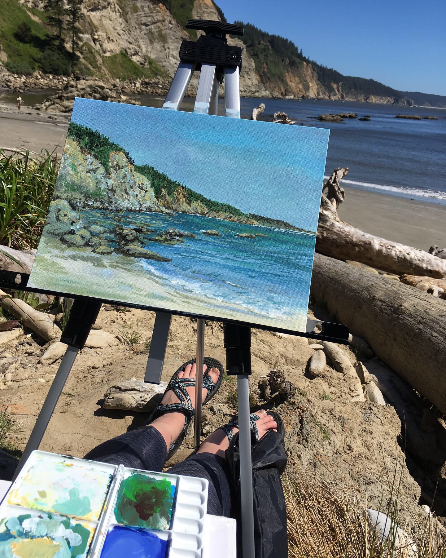Beach scene with cliffs, ocean, and sky, with an easel and painting in the foreground, and a person sitting on the sand.