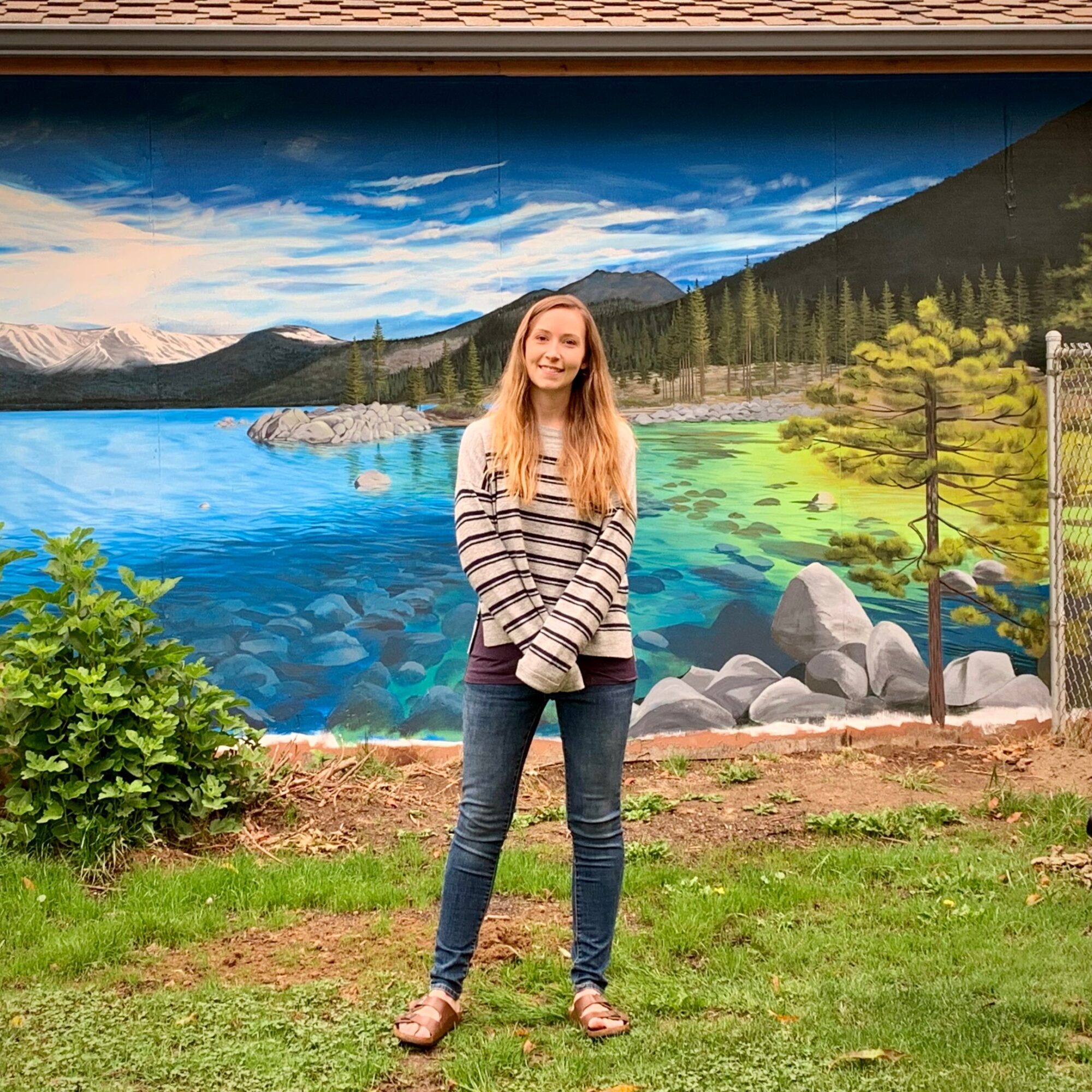 Young woman standing outdoors in front of a painted landscape mural, smiling, wearing a striped sweater and jeans.