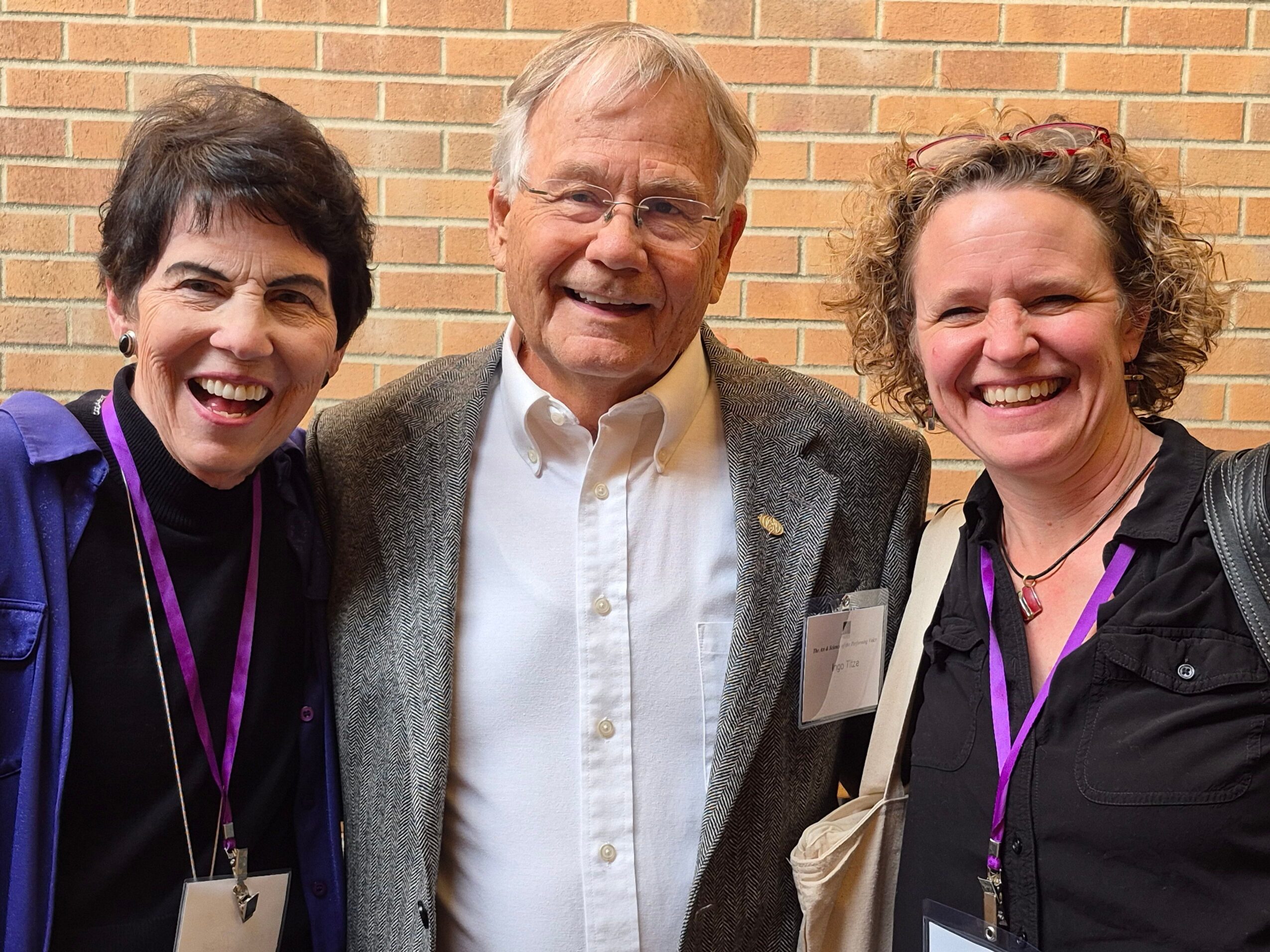 Three people smiling, standing outdoors against a brick wall, wearing casual and semi-formal clothing.