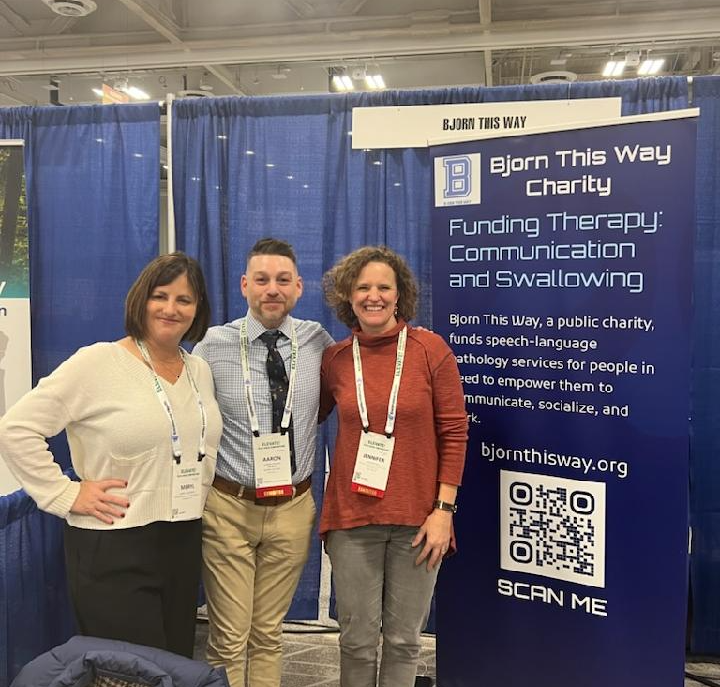 Three people standing in front of a blue charity booth with a sign and QR code, smiling at the camera.