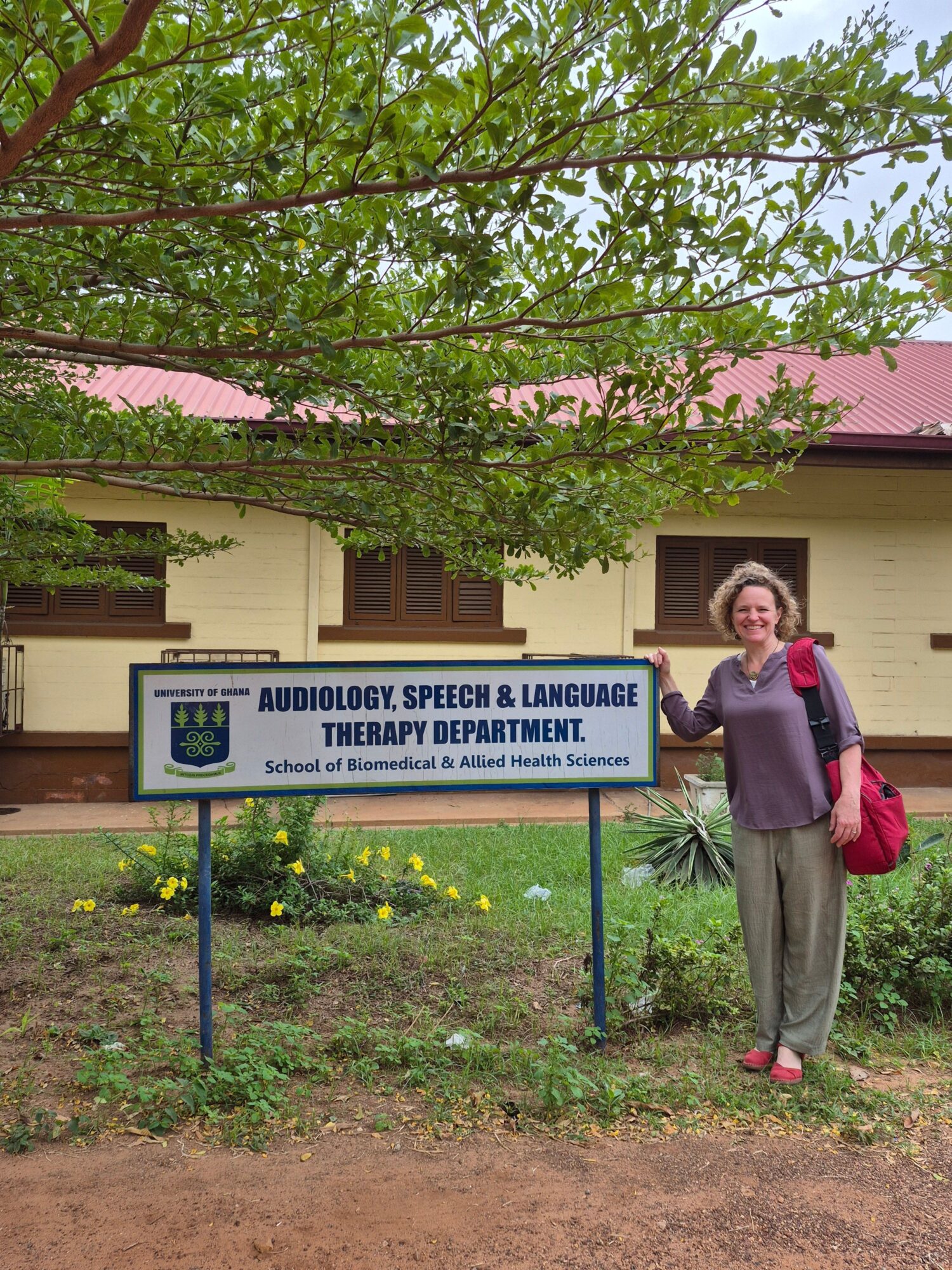 Woman standing outdoors next to a sign in front of a building with trees and grass.