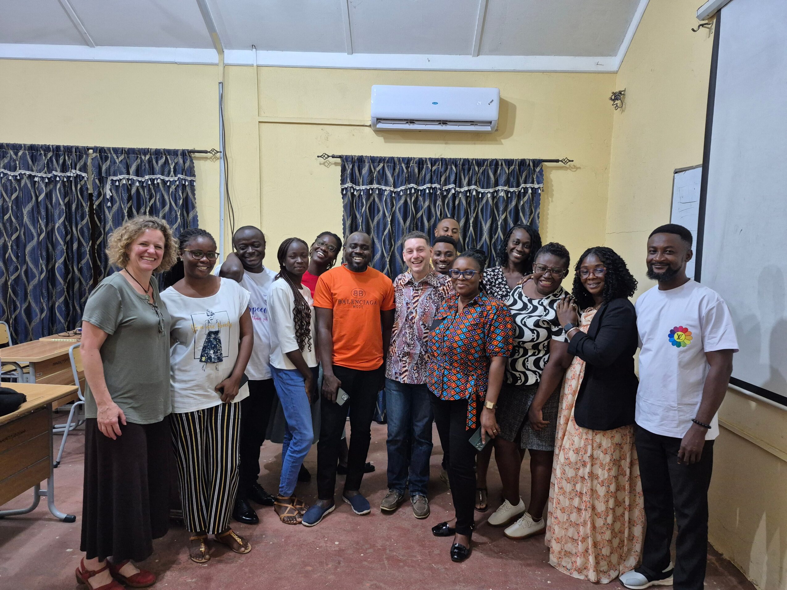 Group of 14 people standing in a classroom with curtains, air conditioner, and whiteboard, smiling for a photo.