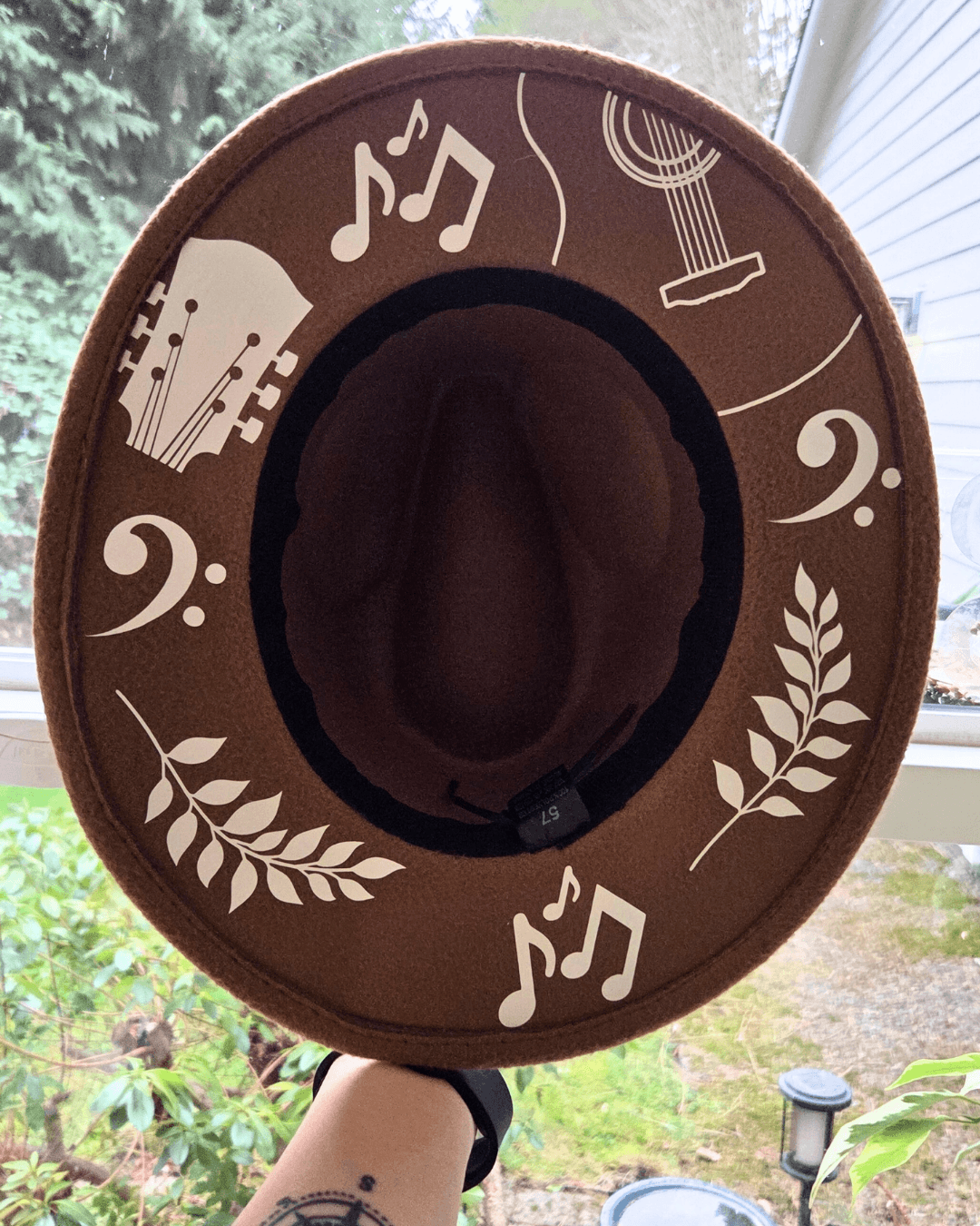 Brown hat with musical notes, guitar, and leaf designs, held outdoors near a window with trees and garden visible.