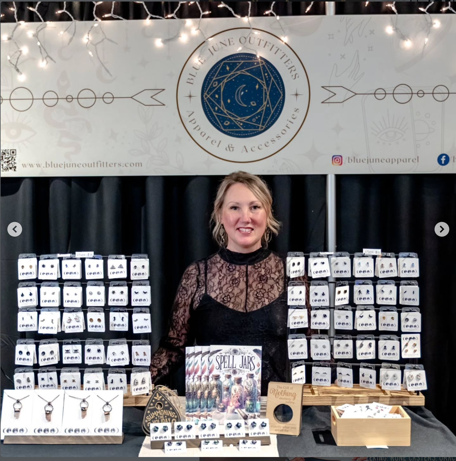 Young woman standing behind a display table with jewelry and a sign, black curtain backdrop, decorative banner above. Wordmark and logo visible.