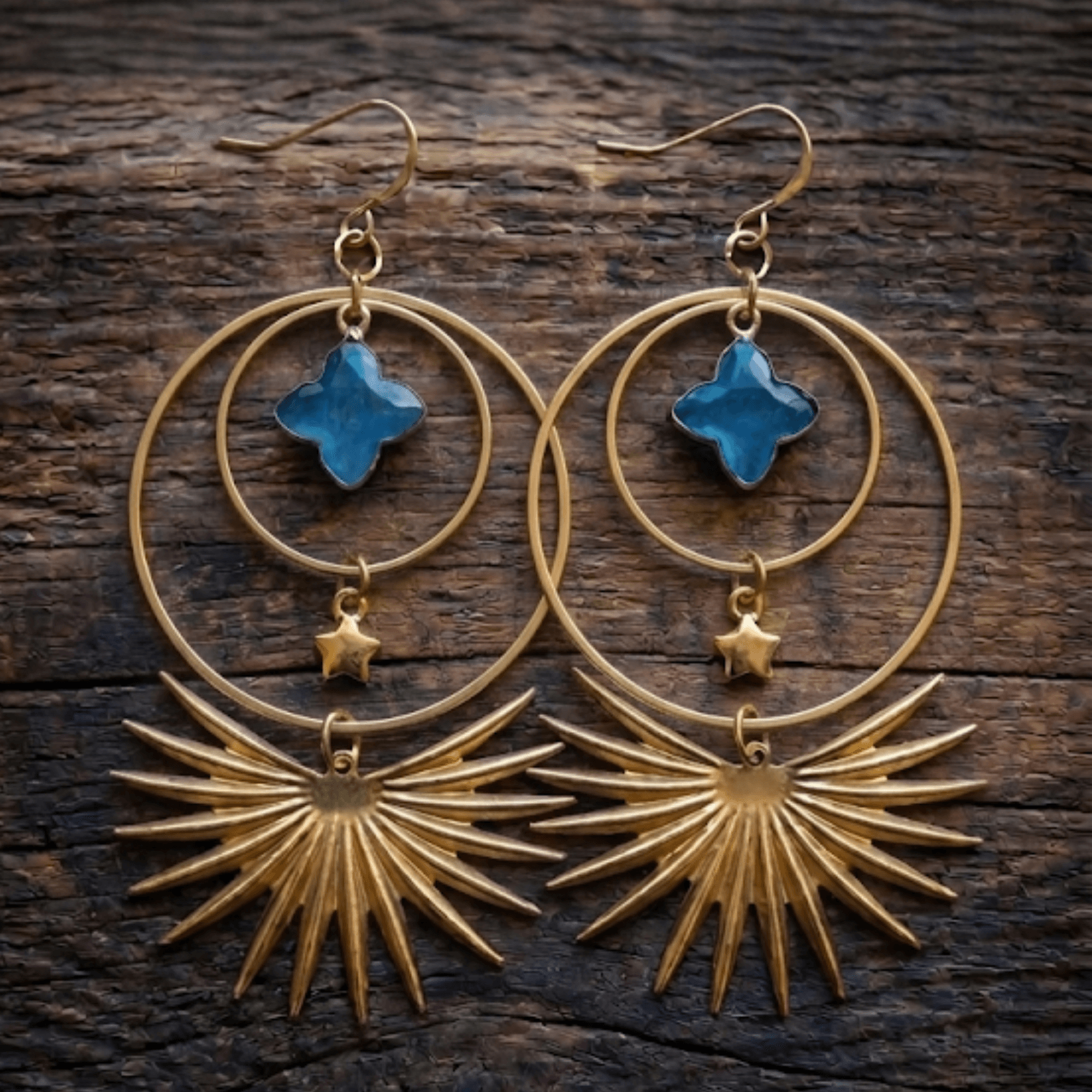 Pair of gold earrings with blue cross-shaped stones, circular and sunburst designs, on a wooden surface.