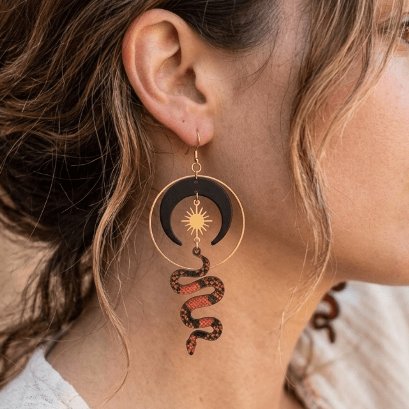 Close-up of a woman's ear with a dangling earring featuring a sun, crescent moon, and snake design.