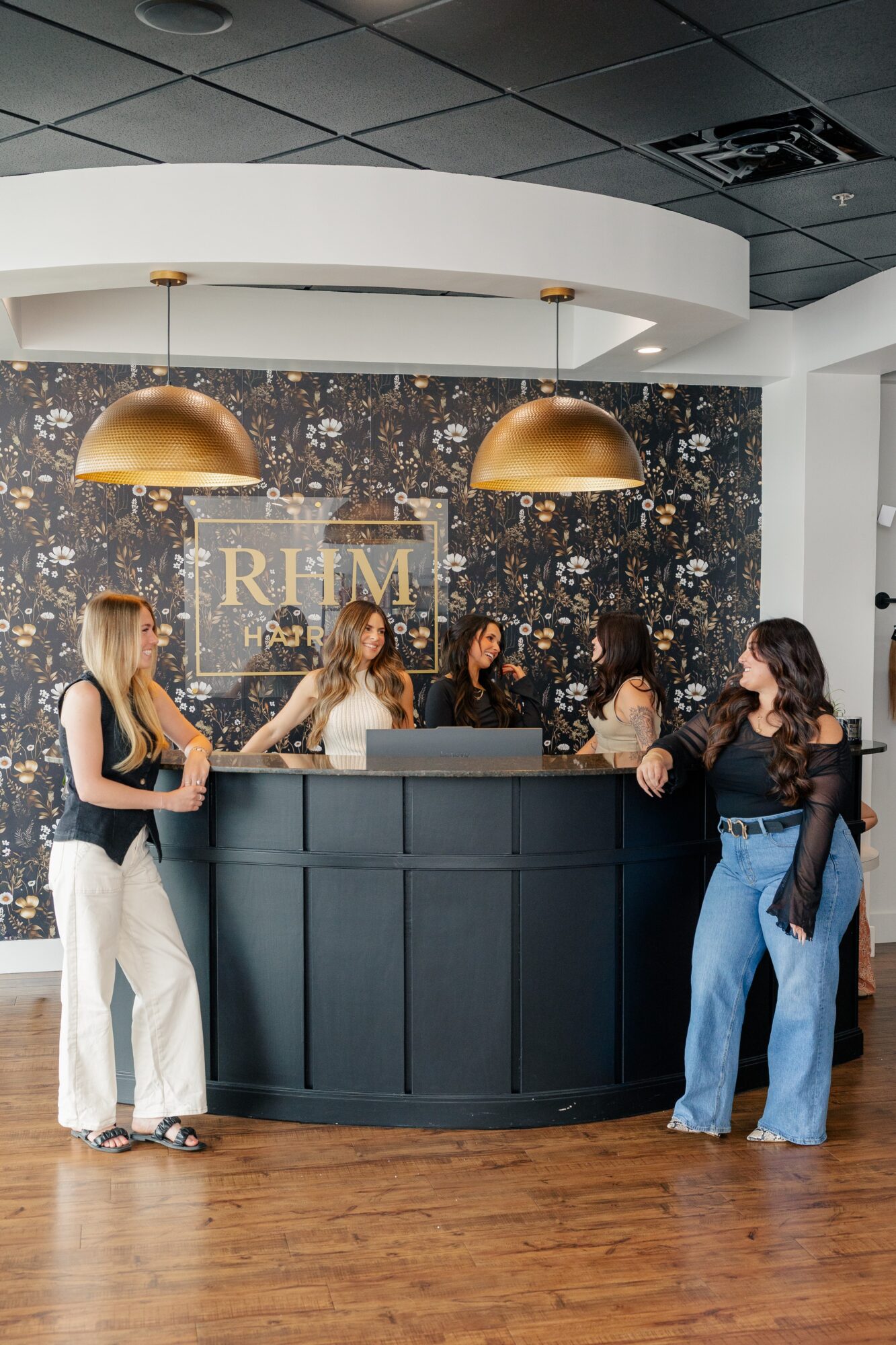 Five women stand at a curved reception desk with a dark front, gold pendant lights, and a textured wall background.