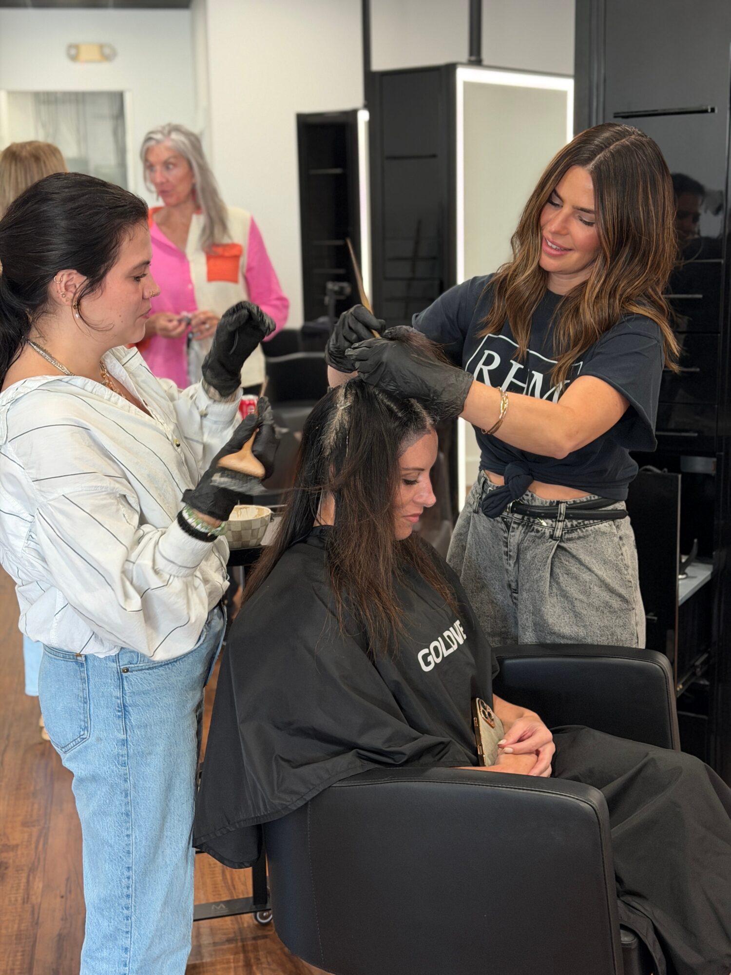 A woman receives a hair treatment from a stylist in a salon, with another stylist working nearby. The seated woman has long dark hair.
