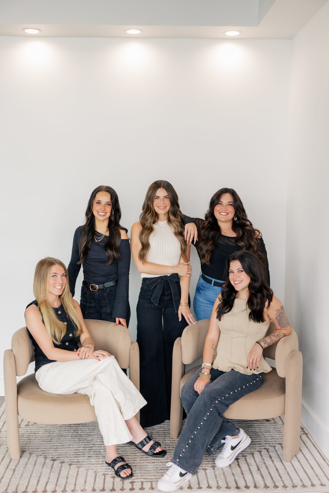 Five women in casual clothing pose in a bright, minimalistic room with white walls and beige chairs.