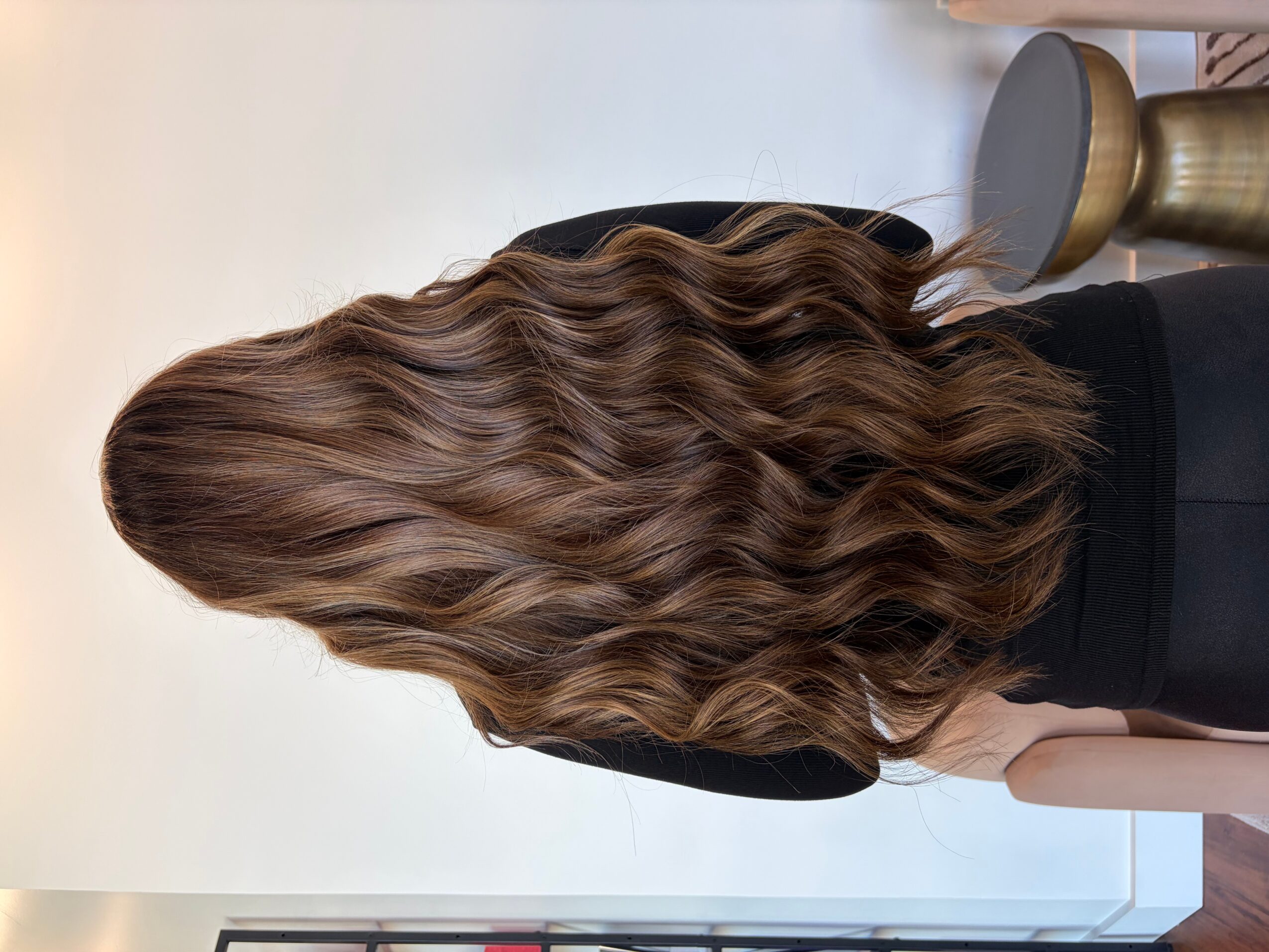 Back view of a woman with long, wavy brown hair, wearing a black top, standing indoors.