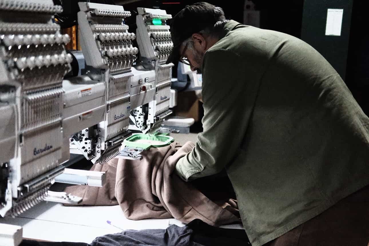 Person sewing fabric on industrial sewing machine in a workshop, wearing glasses and a green jacket, focused on work.